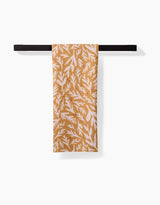 Twig + Sparks Trail Dusting Gold tea towel, featuring a white leafy print and made from recycled materials, hangs neatly over a black towel bar against a plain white background.