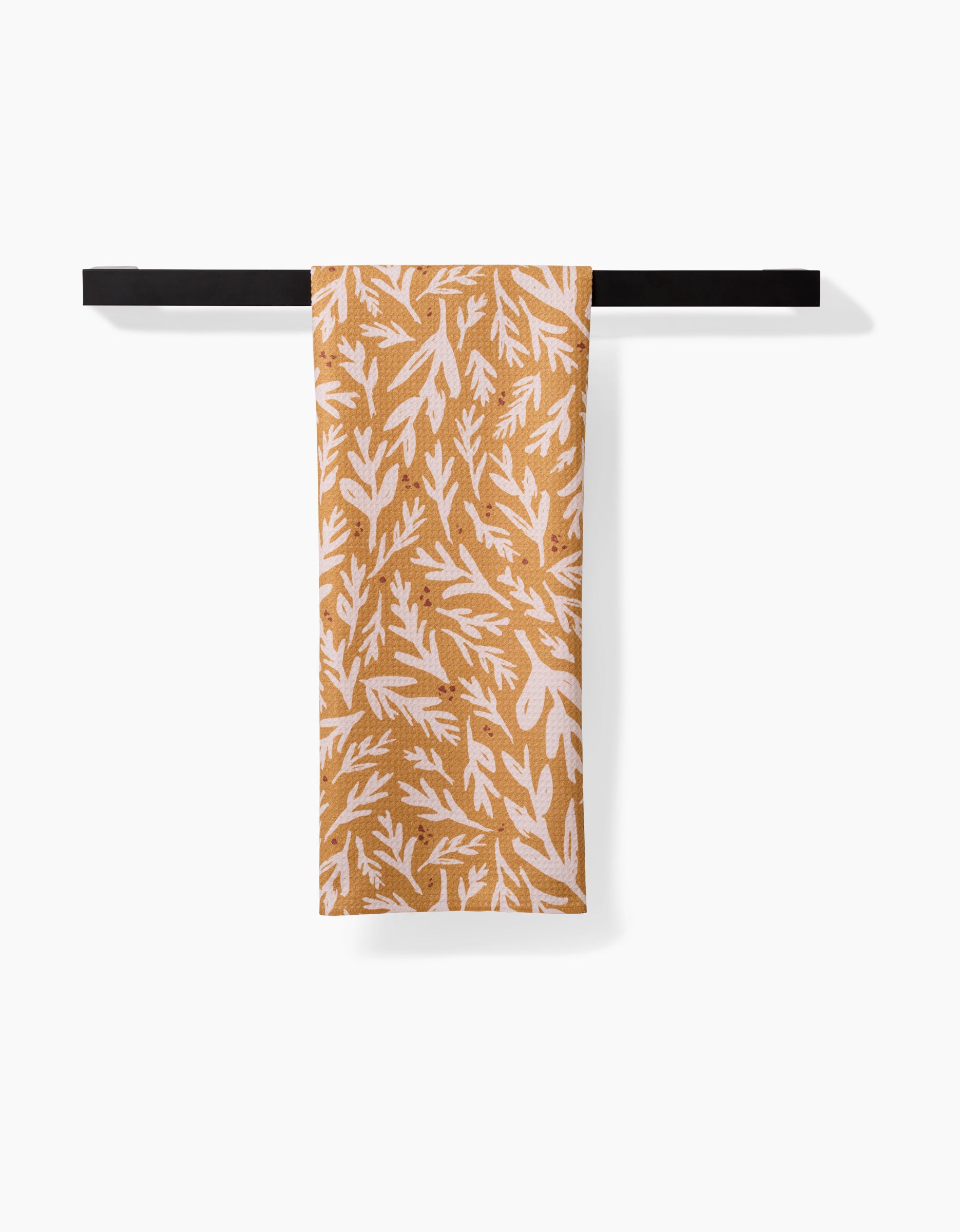 Twig + Sparks Trail Dusting Gold tea towel, featuring a white leafy print and made from recycled materials, hangs neatly over a black towel bar against a plain white background.