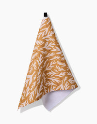 The Twig + Spark Trail Dusting Gold tea towel, ultra-absorbent and featuring a leafy brown and white pattern, hangs from a black loop against a white background, partially folded to show its contrasting white underside.