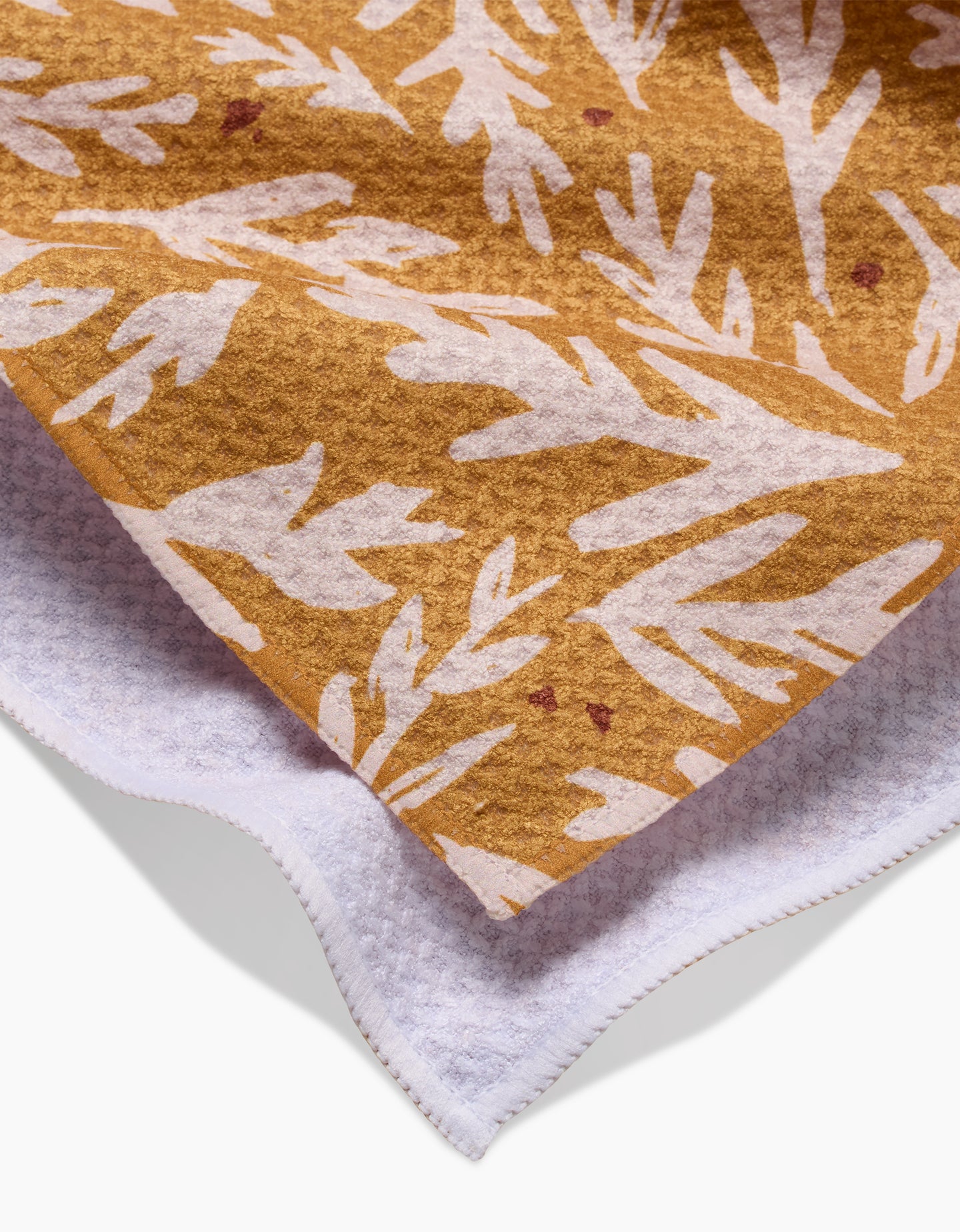 A close-up of Twig + Spark’s Trail Dusting Gold shows its soft, mustard-yellow fabric with white abstract leaf patterns, layered over an ultra-absorbent tea towel for a cozy, textured appearance at the corner.