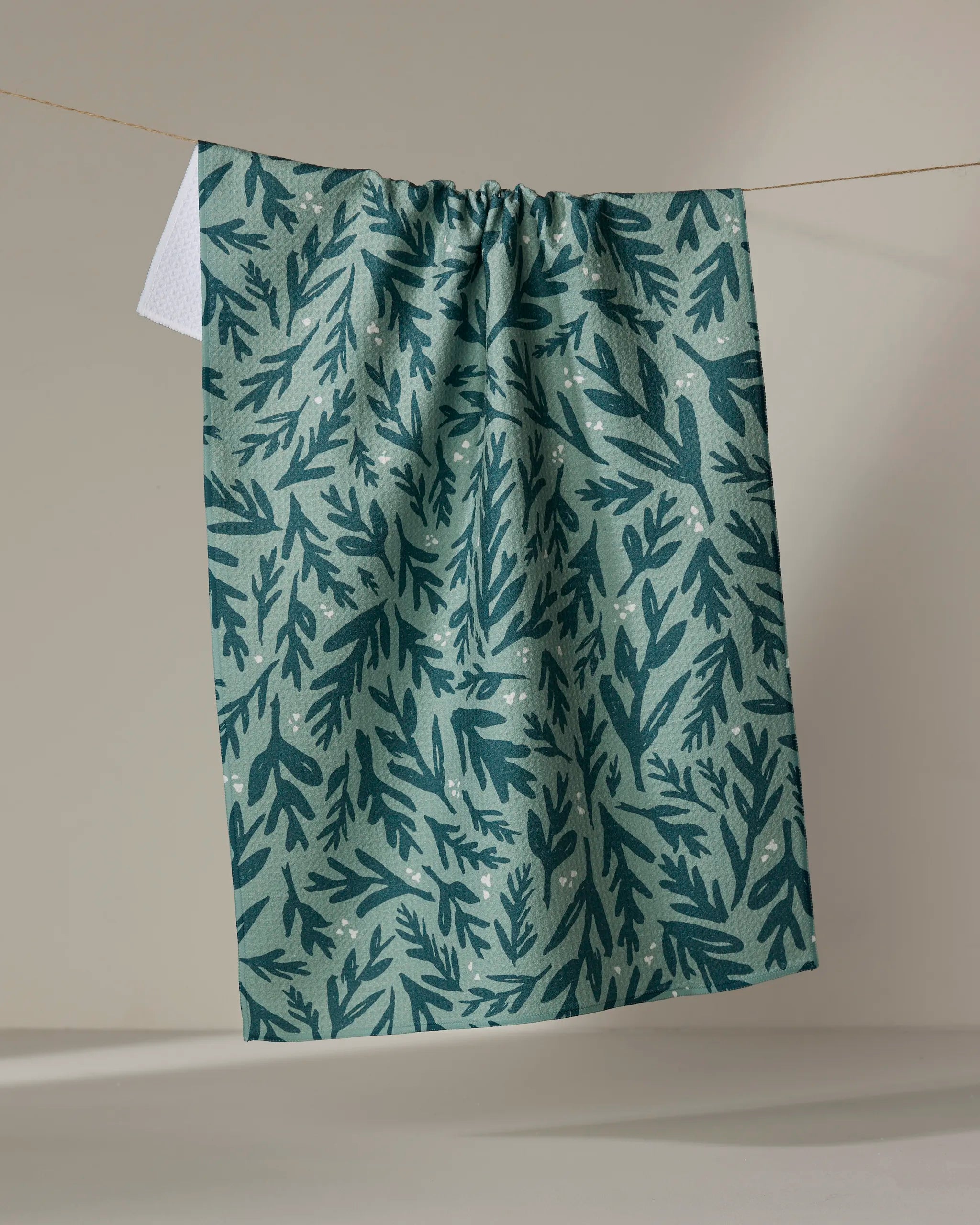 The Twig + Spark "Trail Dusting" green tea towel, featuring a dark green leafy pattern, hangs on a clothesline against a neutral background.