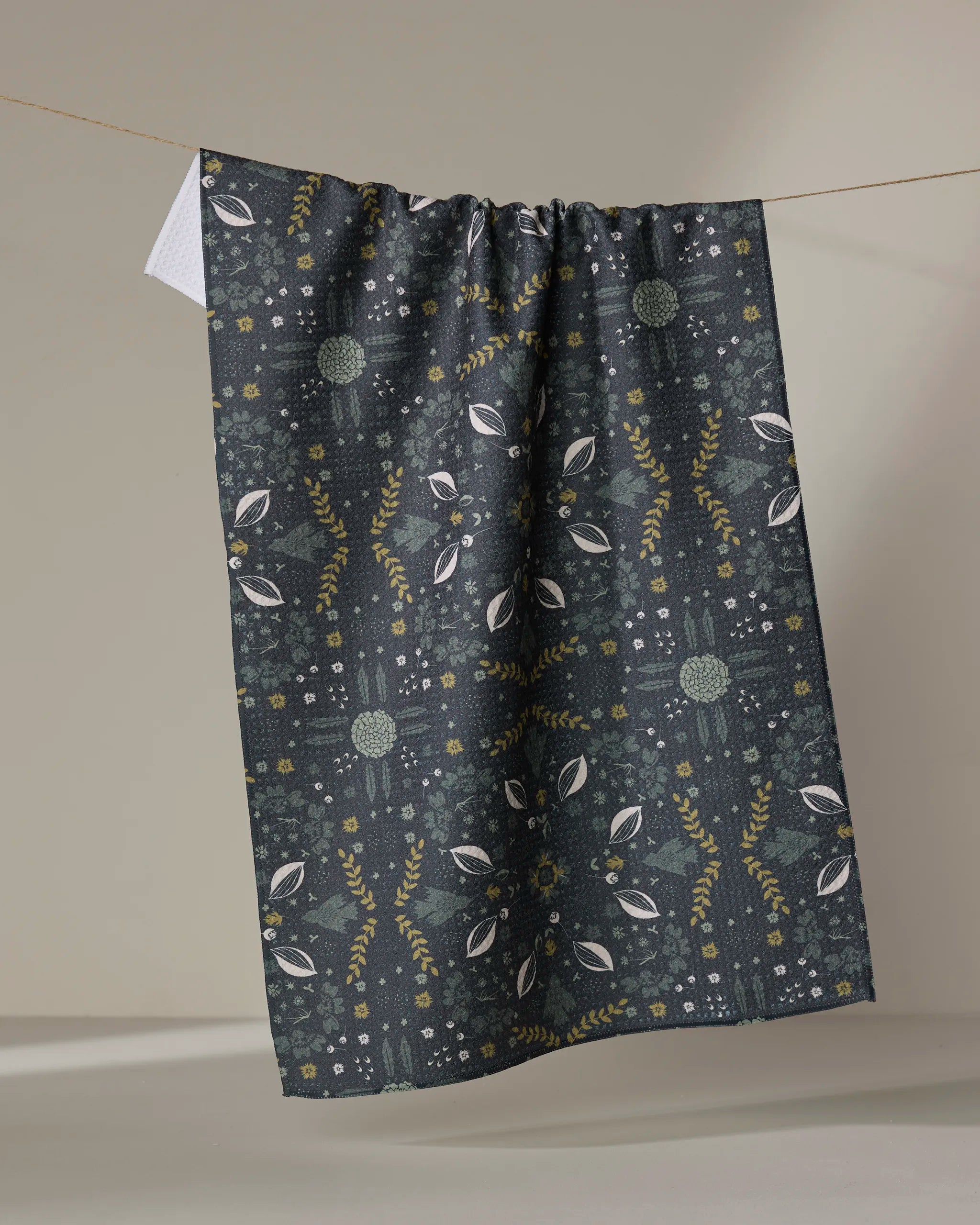 The Ash Cascade Trailhead recycled kitchen towel features a dark base with green, white, and yellow symmetrical floral and leaf patterns and hangs on a string against a neutral background.