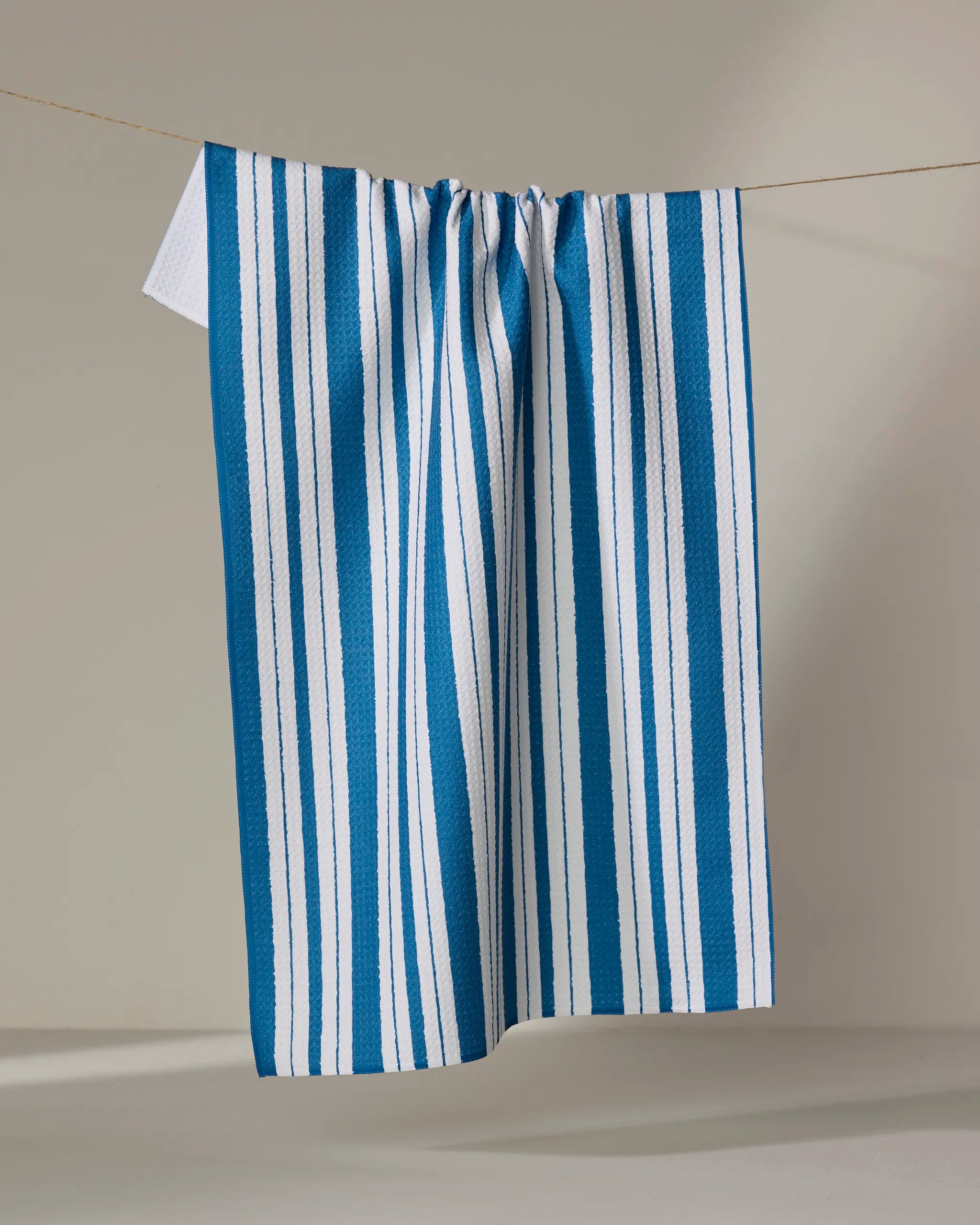 The "Trails of Promise" kitchen towel by Ashley Celena, featuring blue and white stripes, hangs on a thin string against a neutral background for a fresh, simple display.