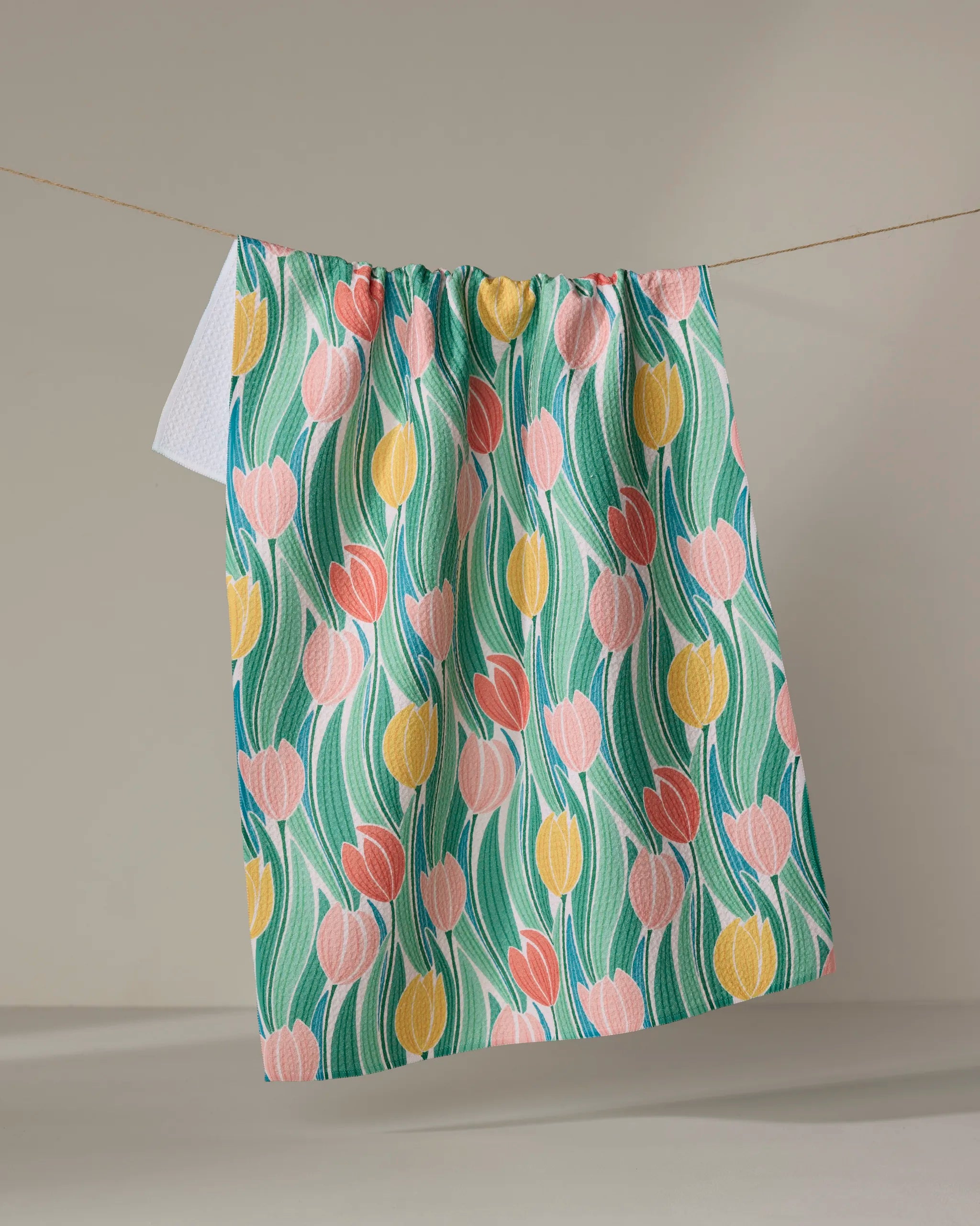 The Tranquil Tulips tea towel by Sarah Daugherty, made from recycled materials and adorned with pink, yellow, and coral tulip patterns and green leaves, hangs on a clothesline against a neutral background.