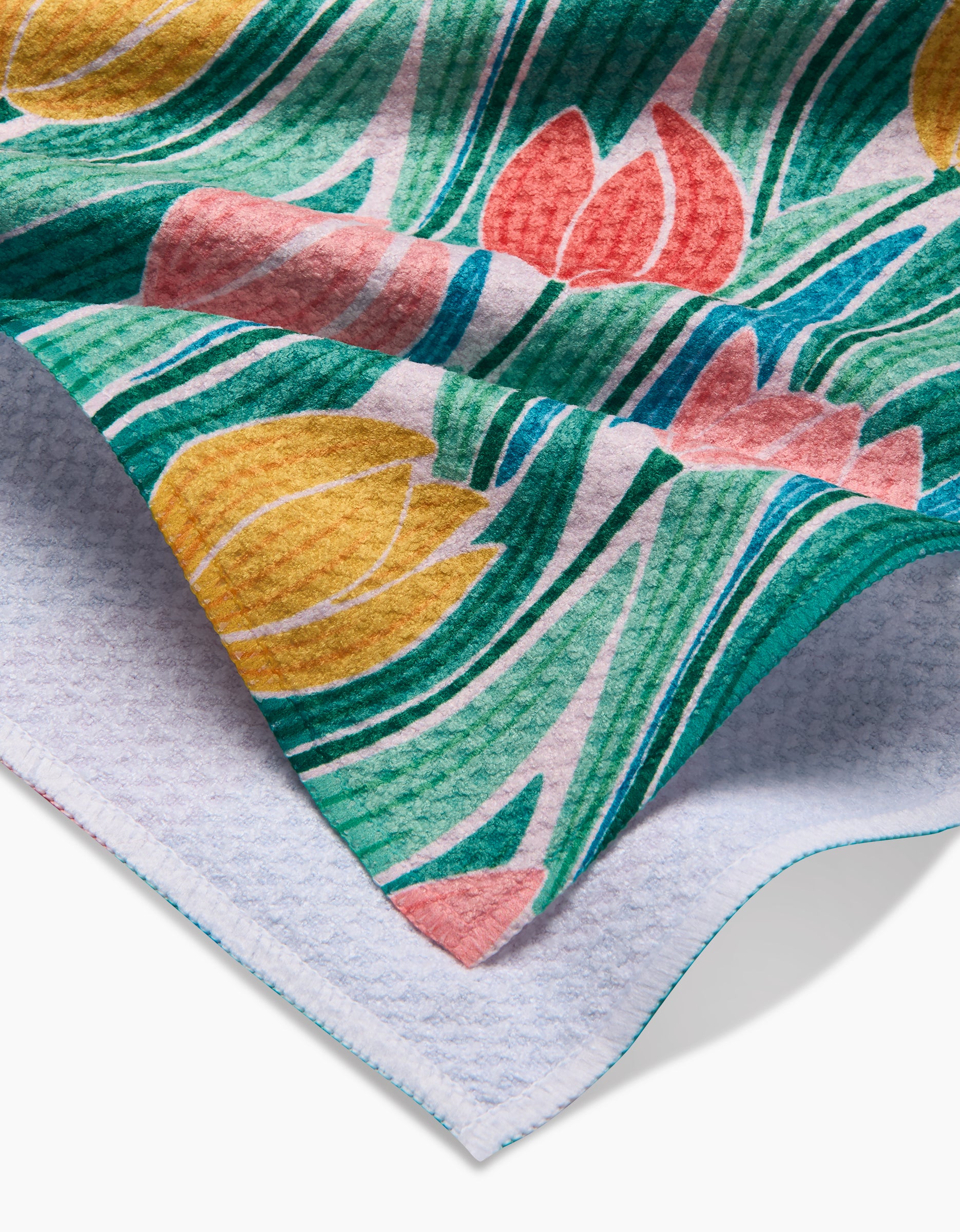 A close-up of the Tranquil Tulips towel by Sarah Daugherty, featuring Geoweave fabric with a vibrant yellow, pink, and green tulip pattern on one side, a white textured reverse, and blue-stitched edges.