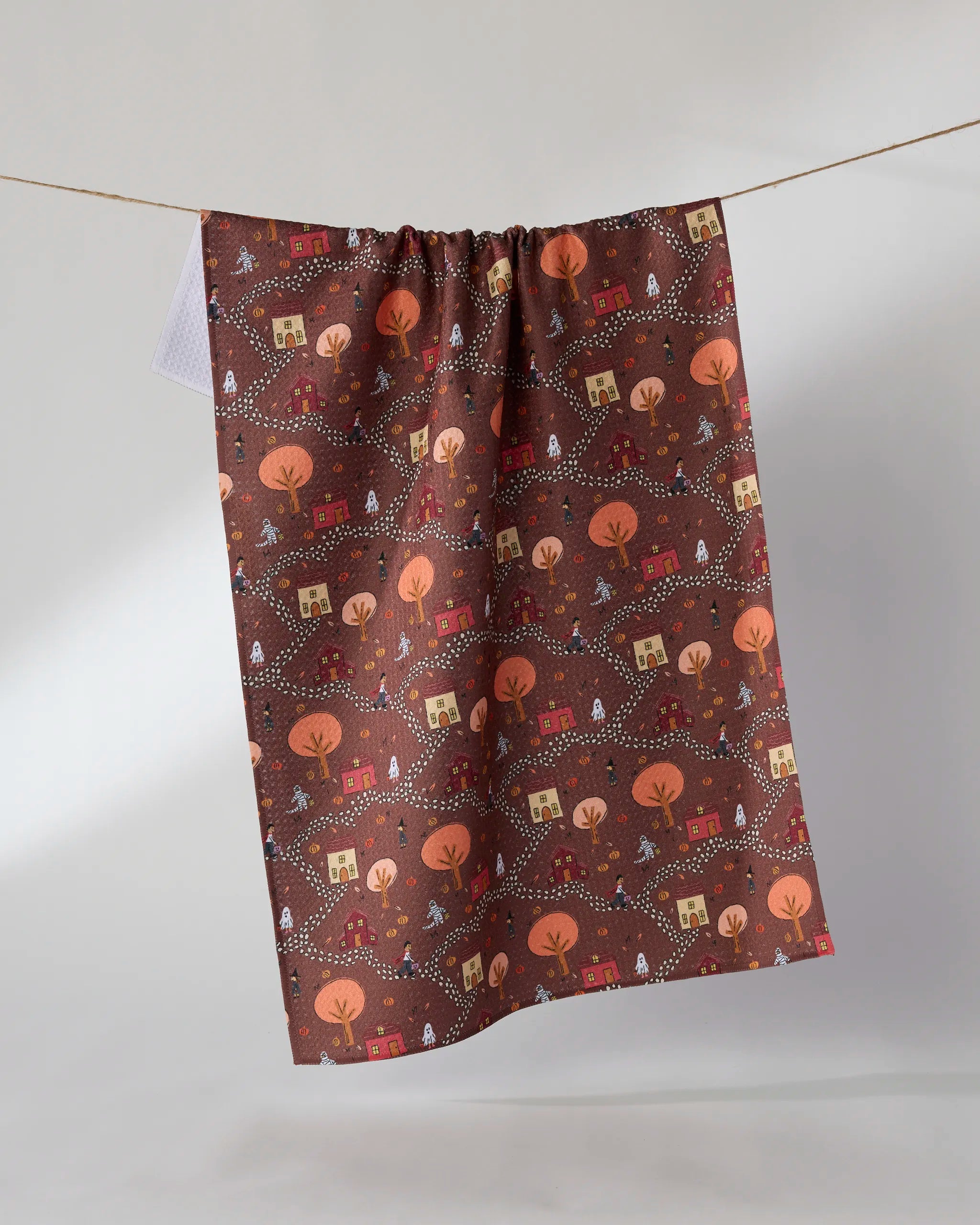 The Geometry Treat Trail kitchen towel features whimsical houses, trees, and winding paths in autumn hues. Made from recycled materials, it hangs on a string against a light backdrop.