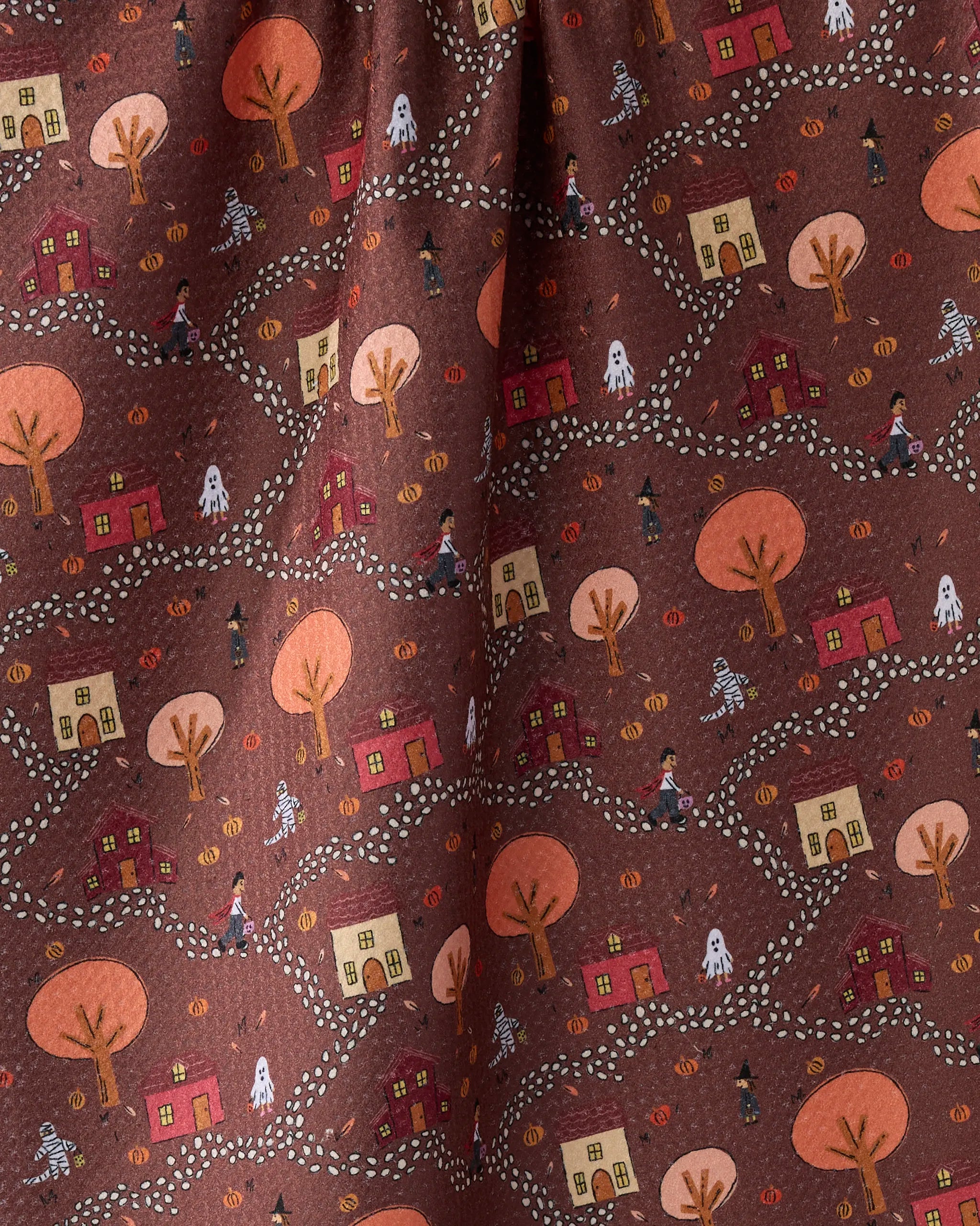 The Geometry Treat Trail tea towel brings festive charm to your kitchen with its Halloween-themed pattern—small houses, bare trees, ghosts, pumpkins, trick-or-treaters, and winding dotted paths on a brown background.