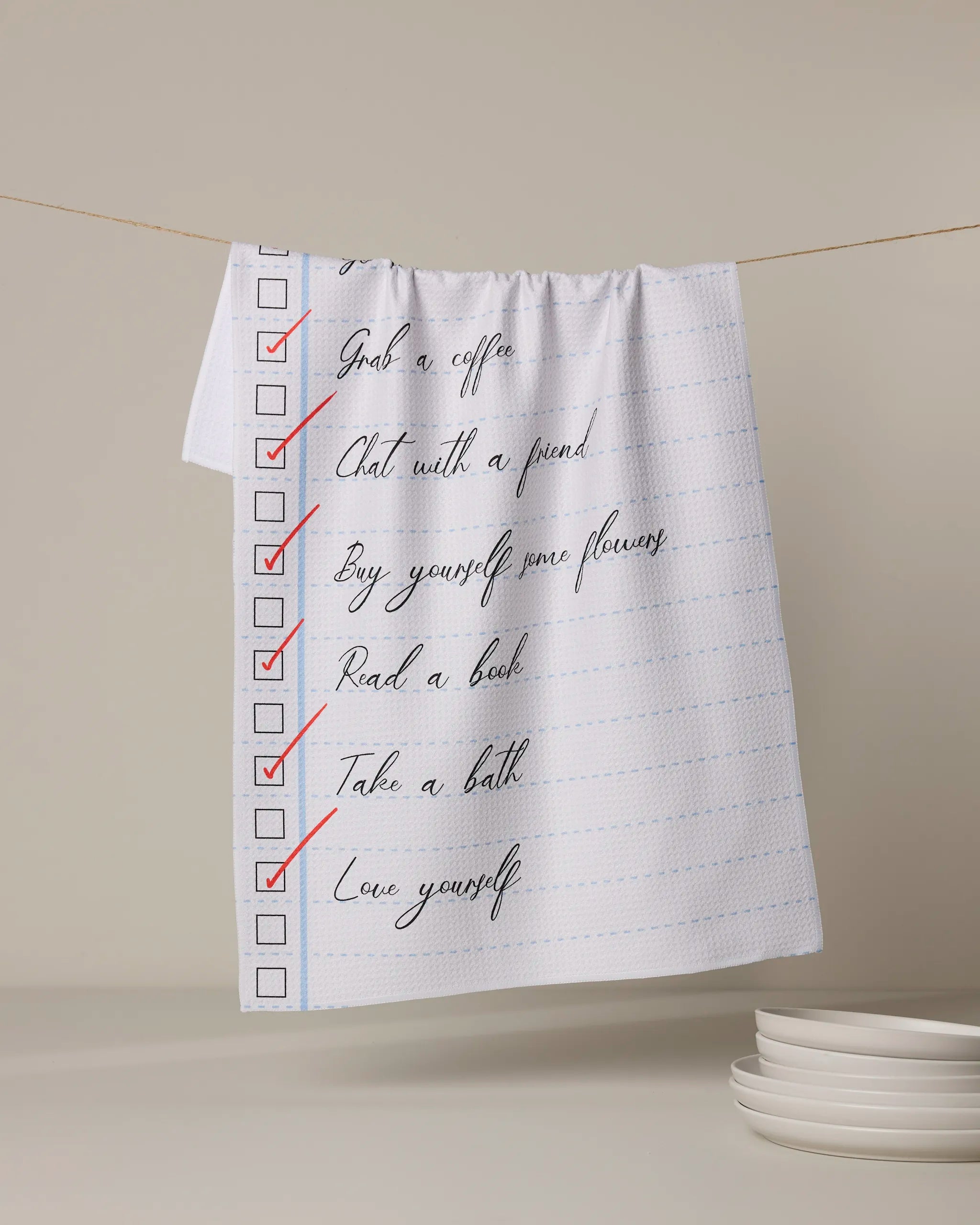 The Treat Yourself towel by Geometry, in white, hangs on a line displaying a red-checked checklist: Grab a coffee, Chat with a friend, Buy yourself some flowers, Read a book, Take a bath, and Love yourself.