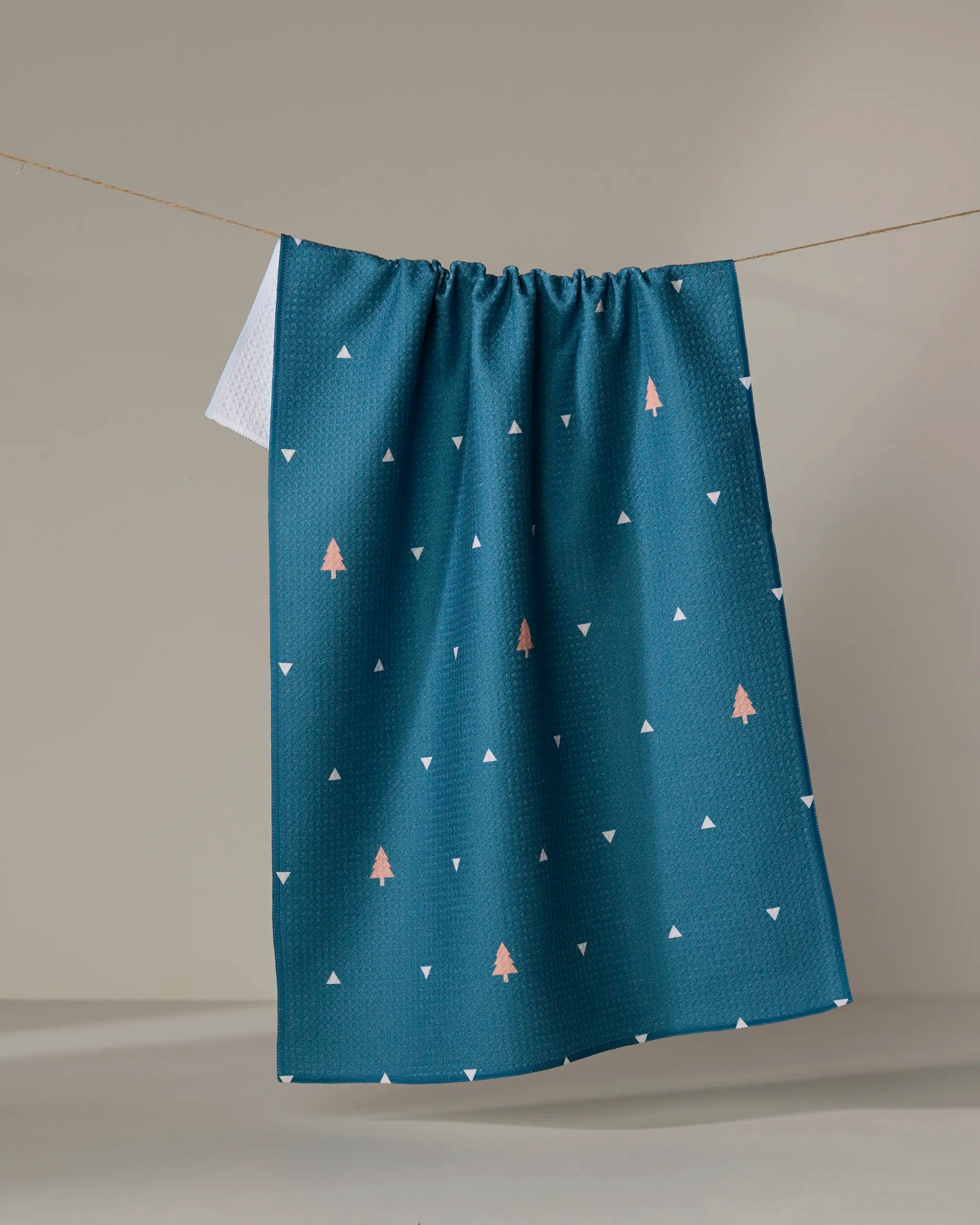 The Twig + Spark Tree Dots tea towel, featuring teal fabric with small white triangles and pink tree accents, made from recycled materials, hangs on a string against a neutral background.