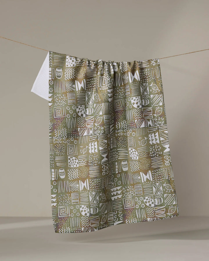 The "Tribal Symbols" tea towel by Taryn Brennan features a geometric abstract pattern in green, brown, and white. Made from recycled materials, it displays unique squares with lines, dots, and shapes against a beige background.
