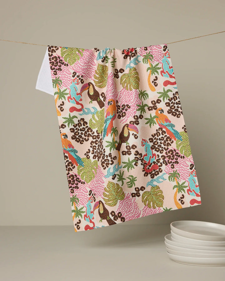 The Geometry Tropi Hotel recycled kitchen towel, featuring a vibrant tropical design with birds, leaves, bananas, and abstract shapes in pink, green, blue, yellow, and brown, hangs above a stack of white plates.