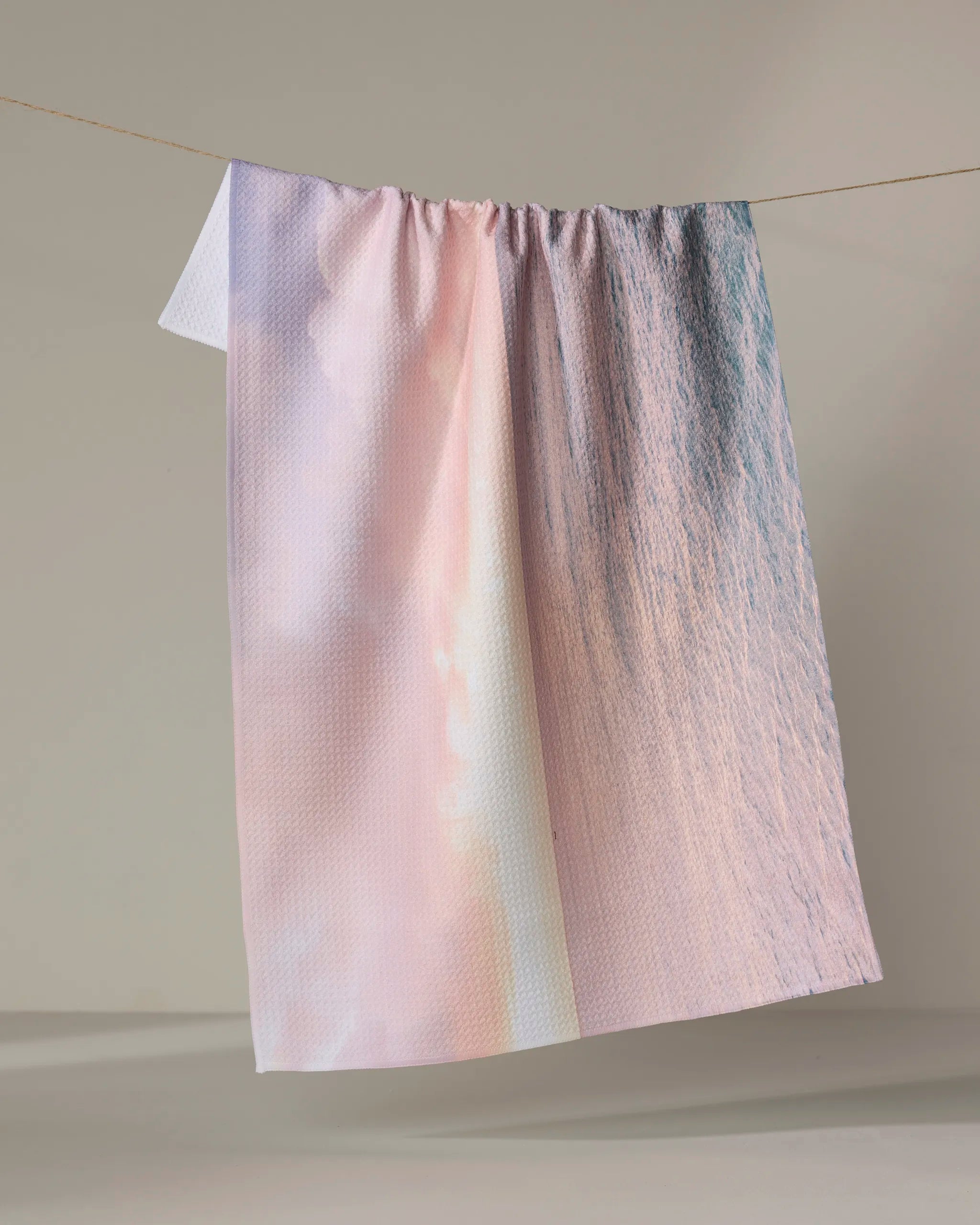 The Kait Hanson "Tropical Sunrise" towel, featuring a pastel gradient of pink, lavender, and blue, hangs textured and absorbent over a clothesline against a neutral backdrop.