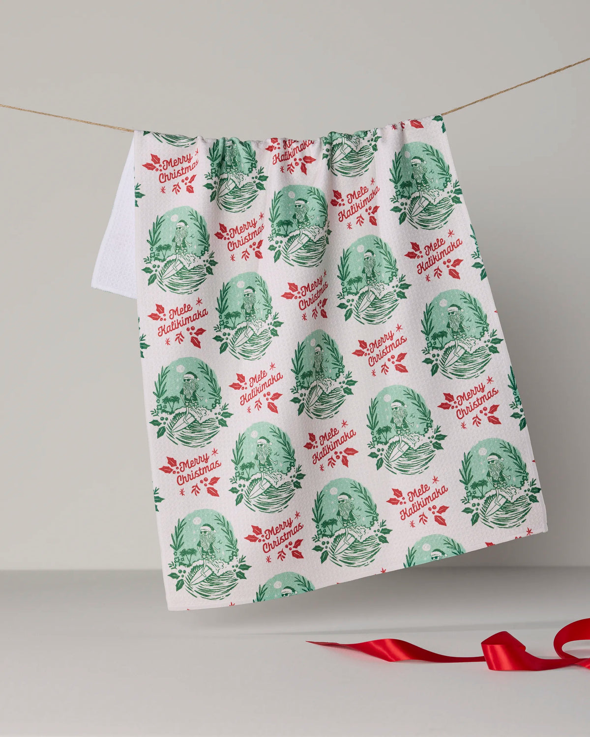 Introducing the Geometry “Tropical Tidings” tea towel, featuring vibrant green and red illustrations and a repeating “Merry Christmas” phrase across its quick-drying fabric. Perfect for the festive season, this towel hangs on a line with a ribbon resting below.
