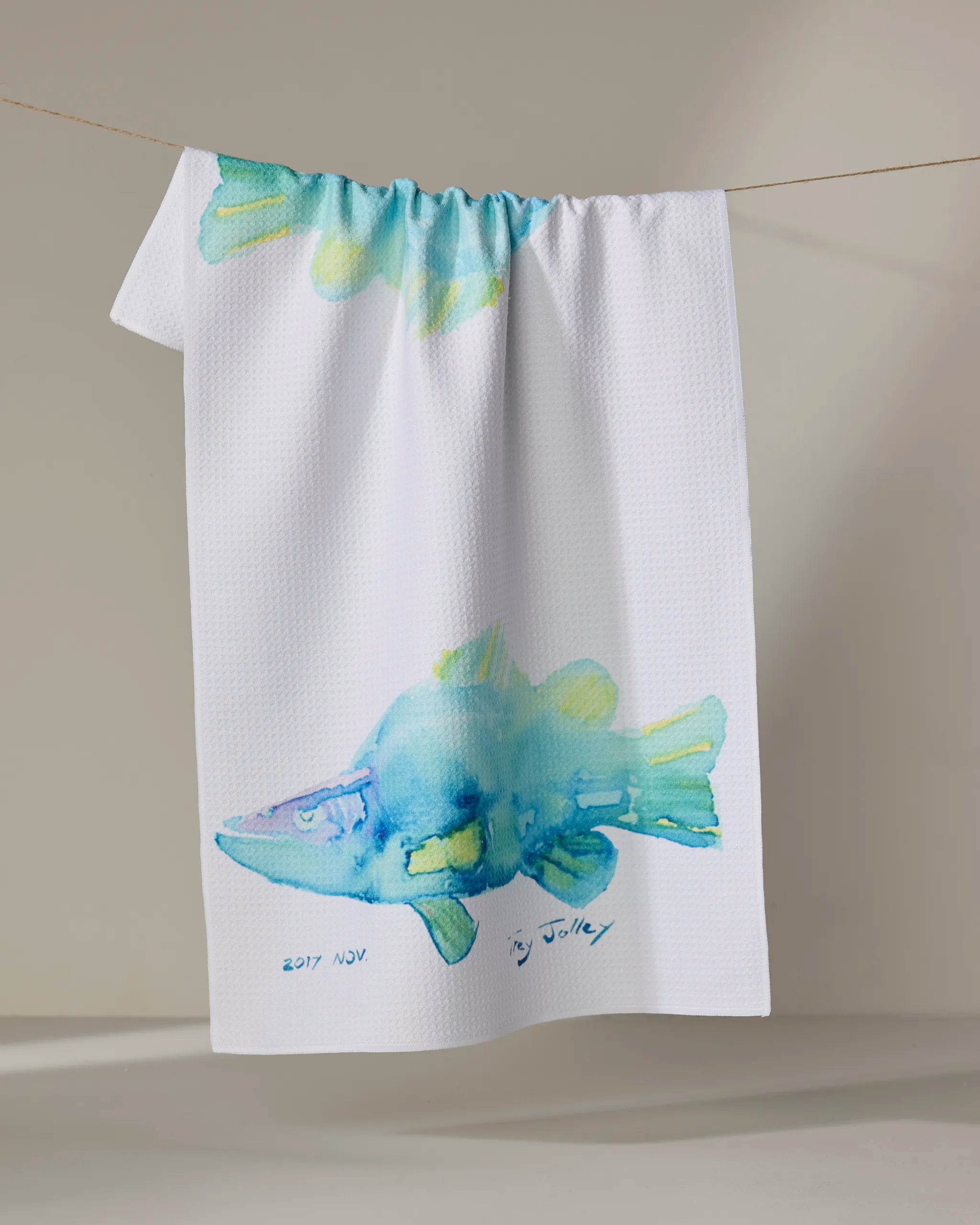 The "Tropical Twins" kitchen towel by Trey Jolley features a watercolor blue and yellow fish on white recycled fabric, hanging on a clothesline against a neutral background. Signed and dated "2017 NOV" at the bottom.