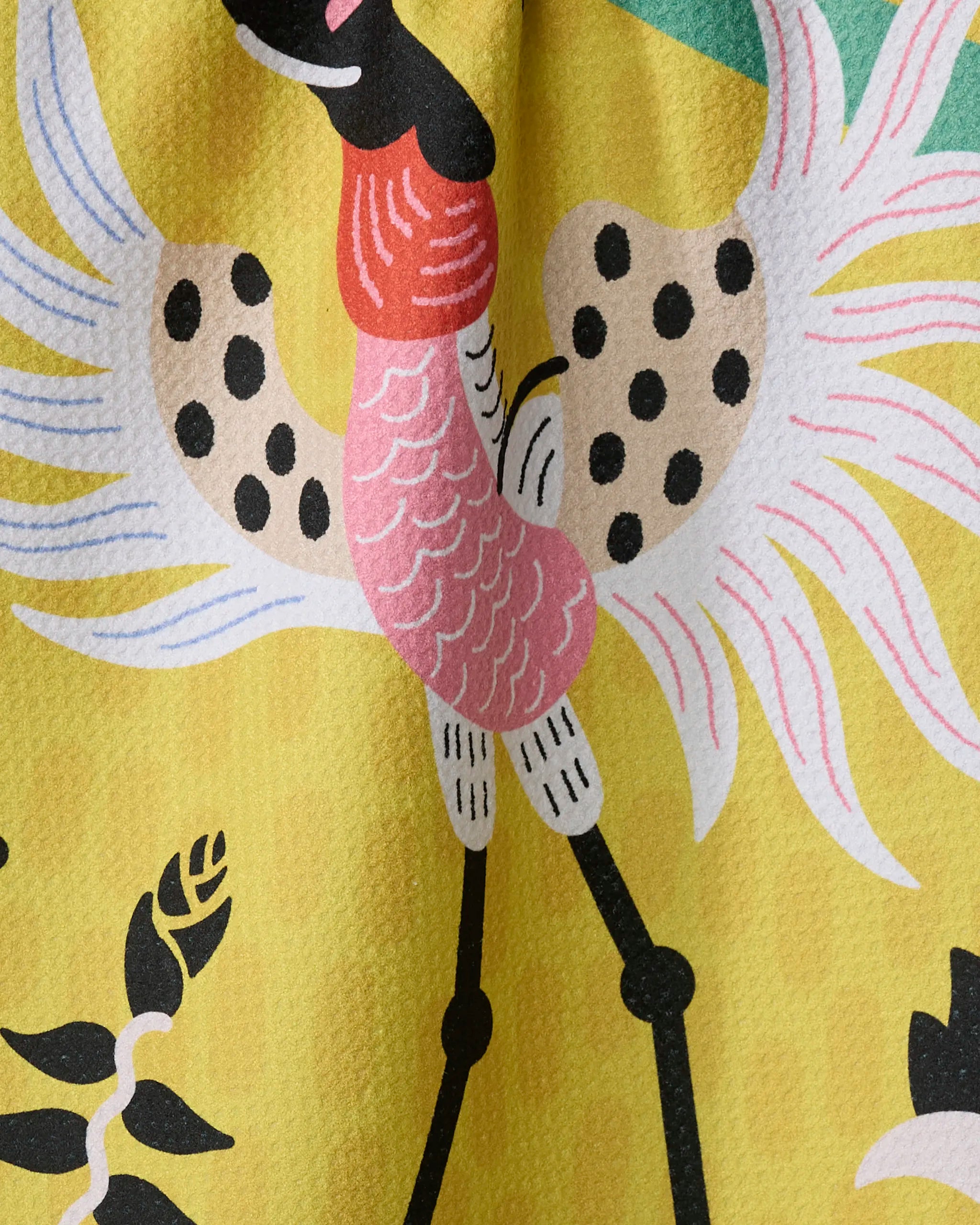 A close-up of Camila Leão’s Tuiuiú shows a stylized bird with a pink neck, black-spotted wings, and long black legs on yellow, surrounded by abstract florals—perfect for a recycled-material tea towel.