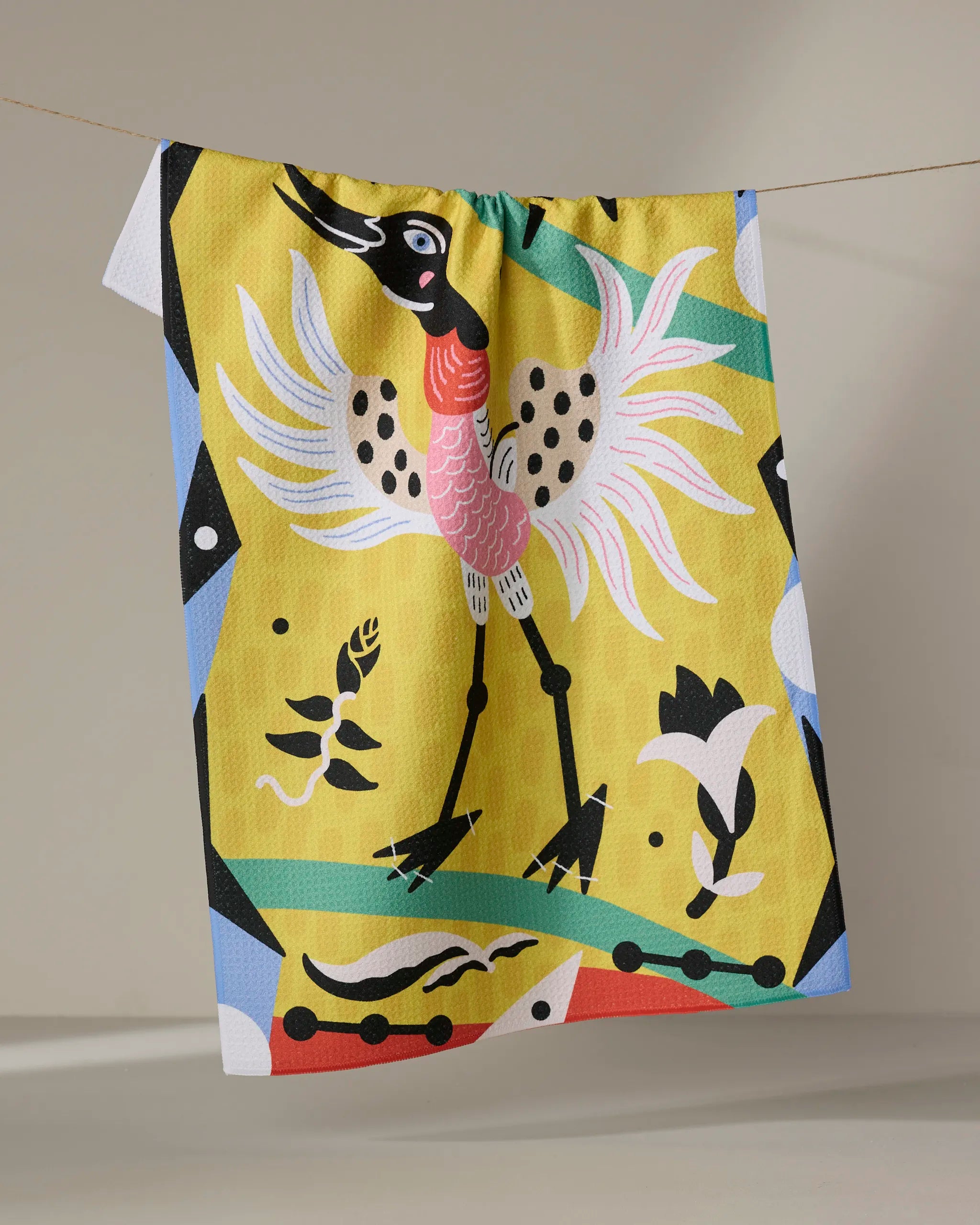 The Tuiuiú Tea Towel by Camila Leão features a stylized crane with a pink chest, black wings, and long legs on a yellow background with bold abstract shapes—crafted from recycled materials.