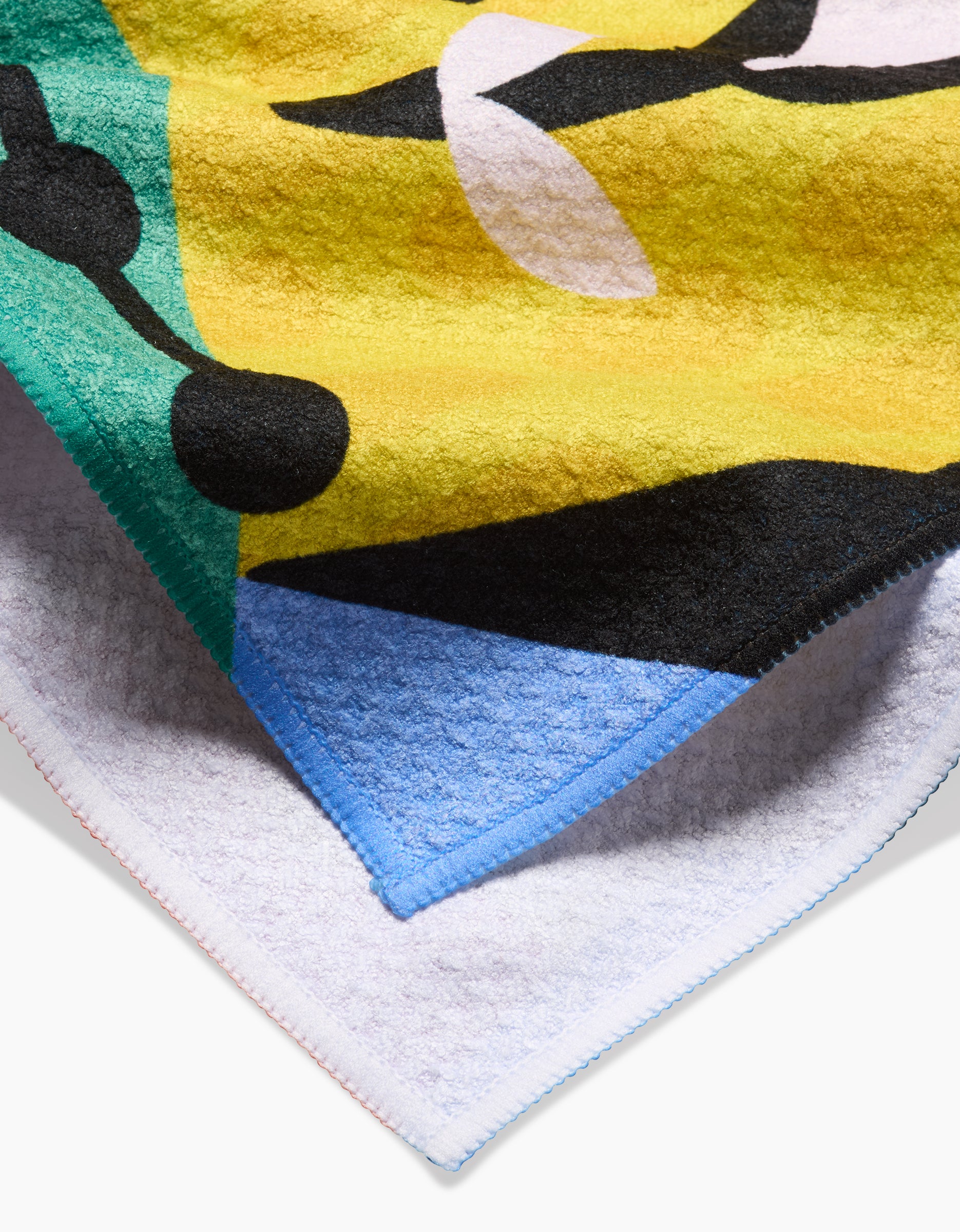A close-up of Camila Leãos Tuiuiú Tea Towel highlights bold abstract shapes in yellow, green, black, blue, and white on textured Geoweave fabric with stitched edges.