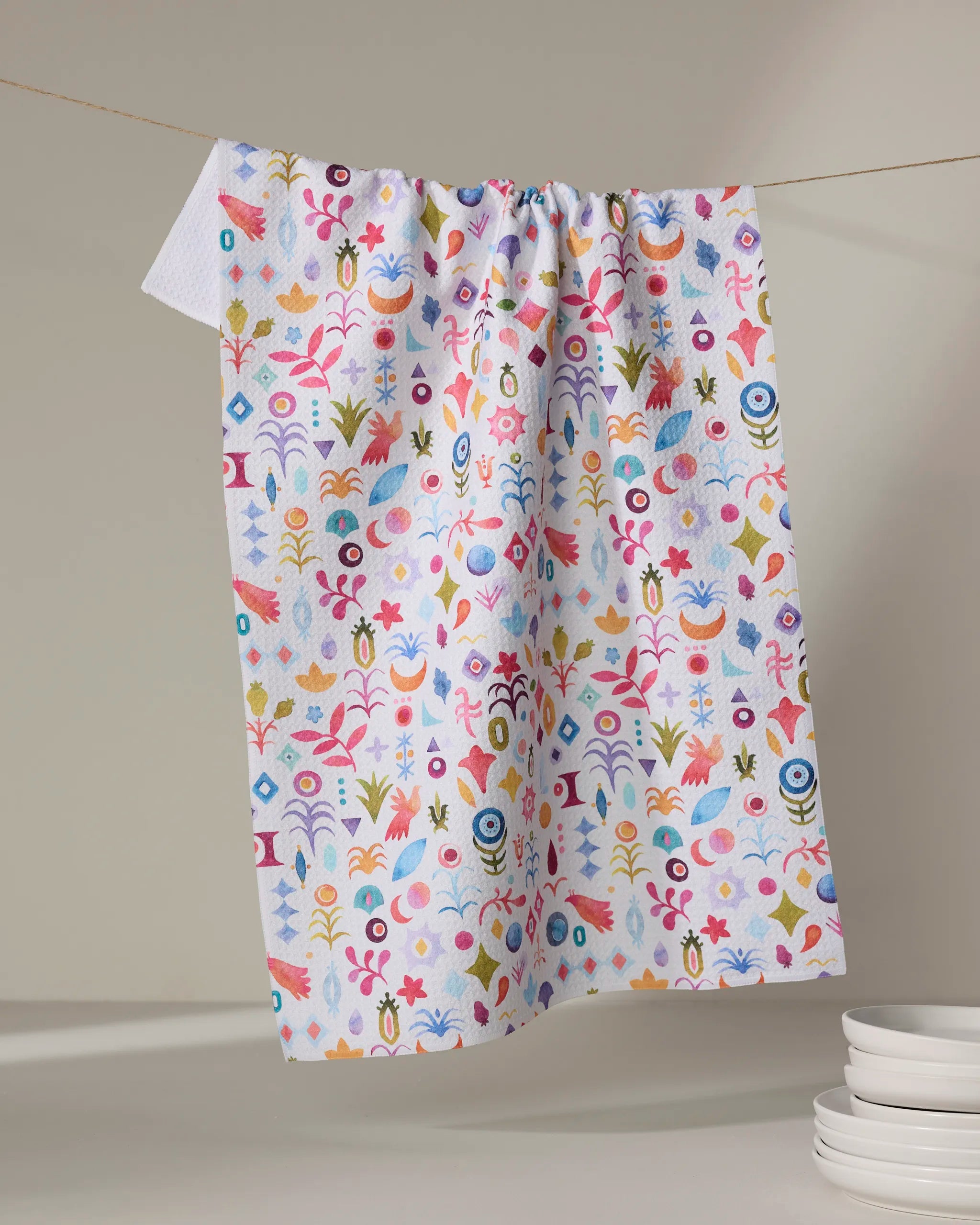 The Tula tea towel by Maayan Alper-Swan, featuring a vibrant floral and geometric pattern, hangs on a line above a stack of white plates. The softly lit background highlights its colorful design.