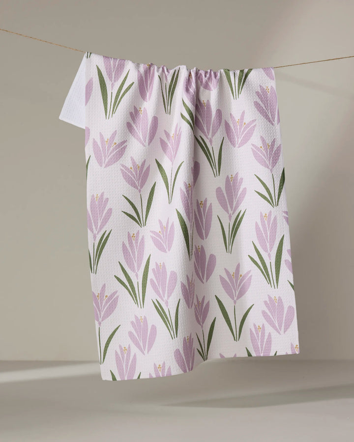 The Tulip Bulbs tea towel by Janna Sue Design features a pattern of light purple flowers and green leaves. Ultra-absorbent and stylish, it’s perfect for adding charm and function to any kitchen décor.