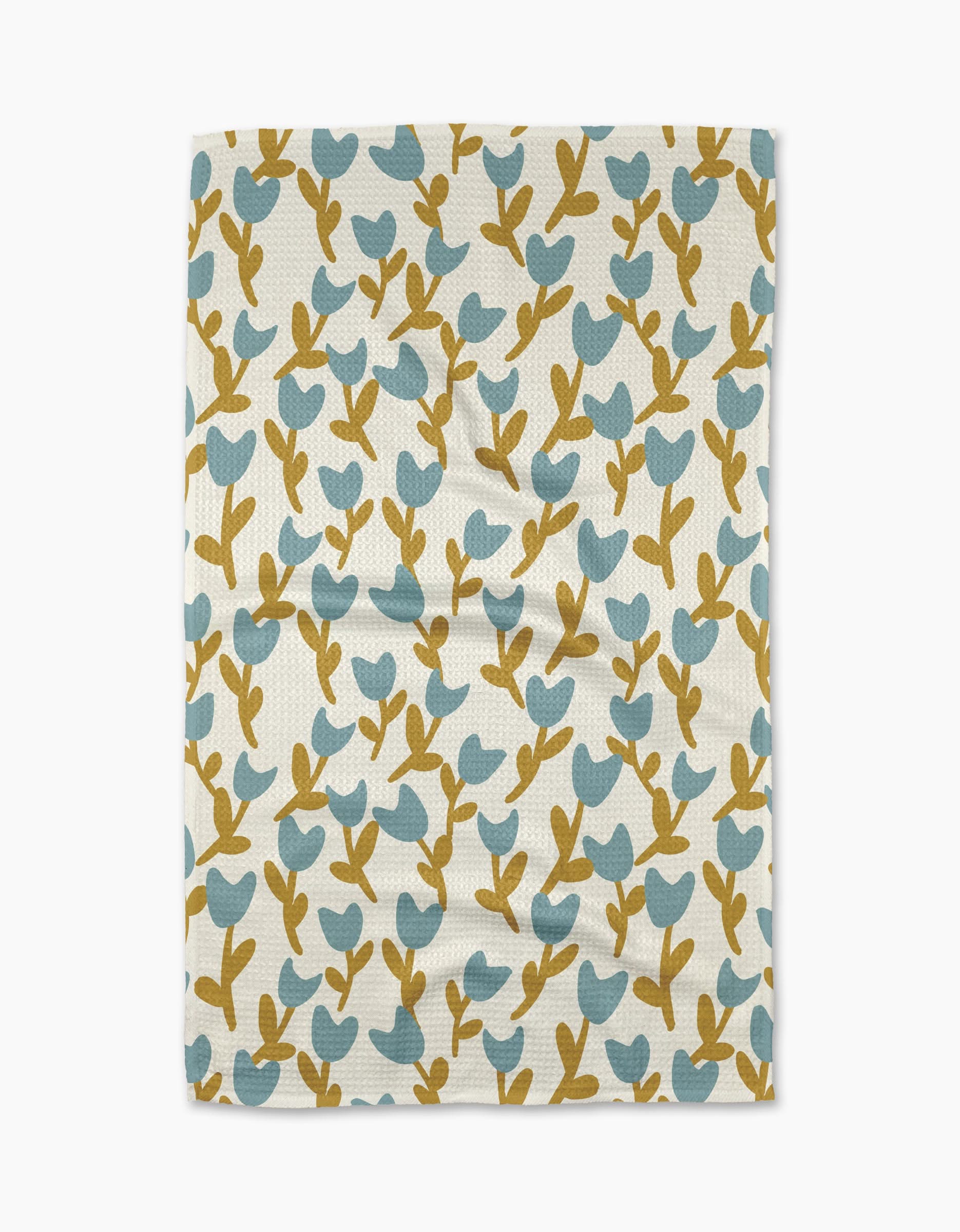 The Tulip Garden tea towel by Lizzie Clark is a rectangular, white towel made from recycled materials, featuring blue and mustard yellow stylized leaf and branch patterns. Displayed flat with slight wrinkles.
