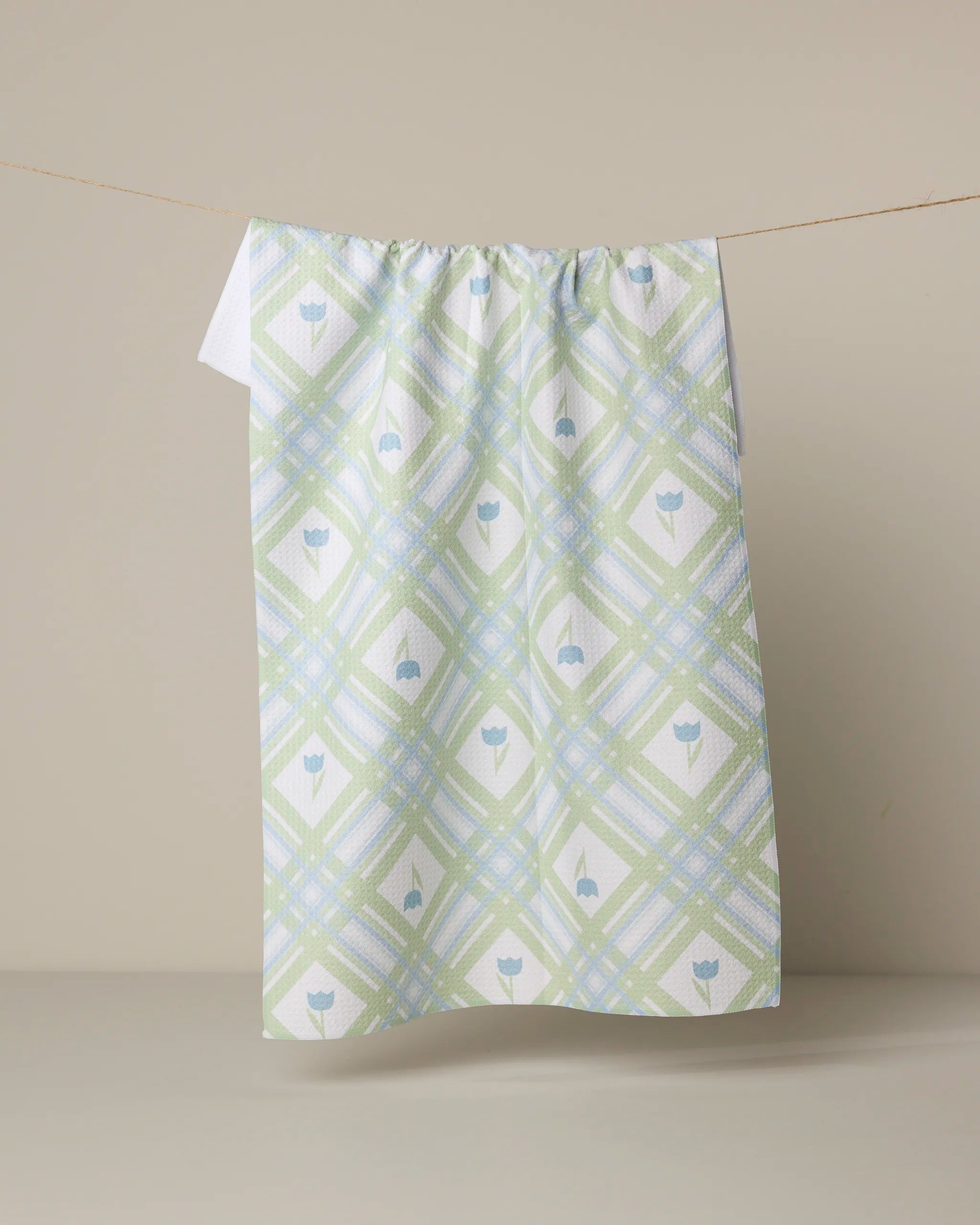 The Geometry Tulip Lattice tea towel features a pastel green, blue, and yellow plaid with blue tulip motifs, made from recycled materials and shown hanging on a clothesline against a beige background.