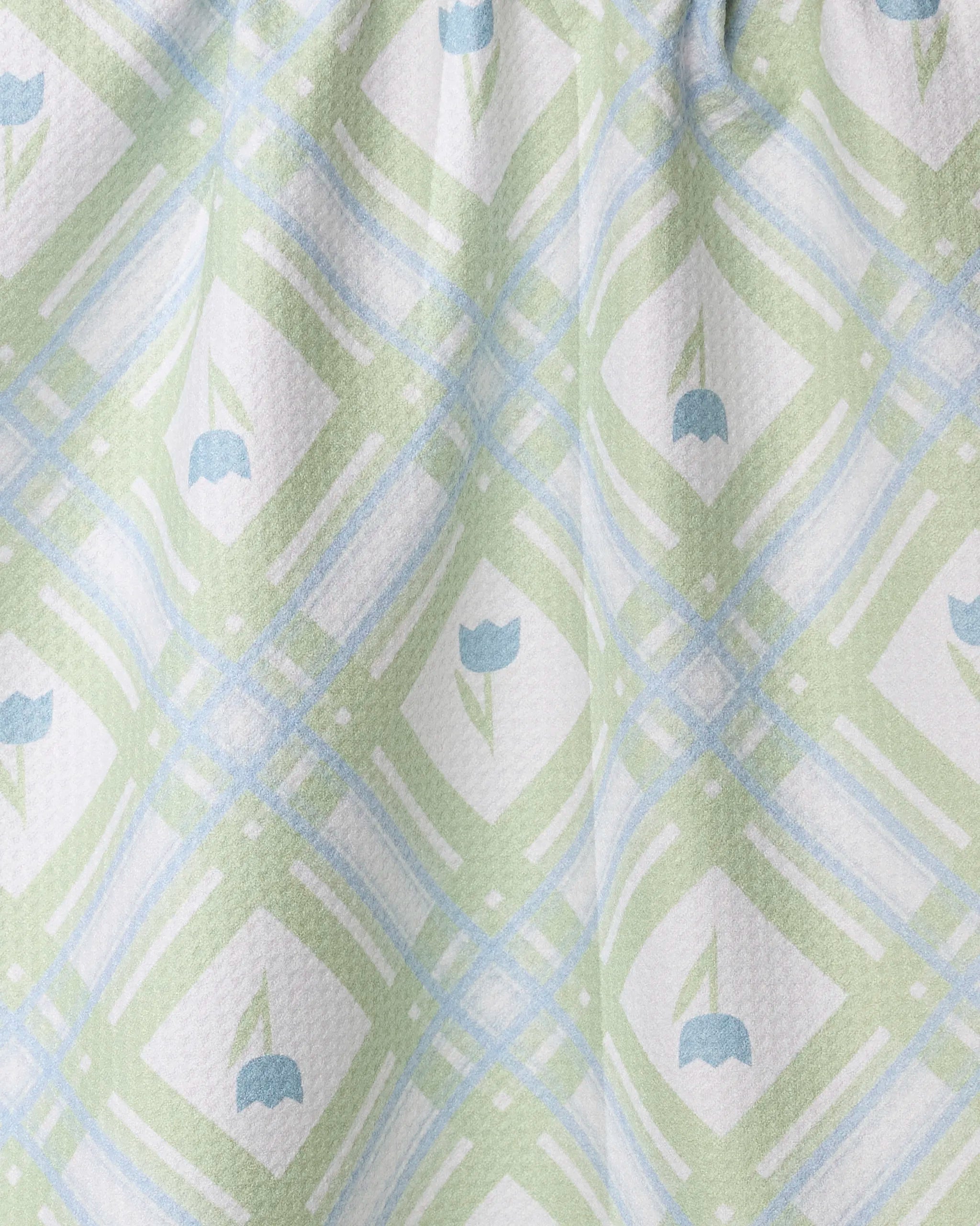 A close-up of Geometry's Tulip Lattice tea towel in pastel green and blue plaid, with white diamond shapes and blue tulip motifs. Made from recycled materials, the ultra-absorbent fabric is textured and gathered at the top.