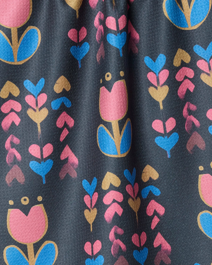 The Geometry Tulip Patch tea towel features a playful, geometric floral pattern with pink, blue, and yellow flowers and leaves in vertical rows on dark fabric, crafted from recycled materials.