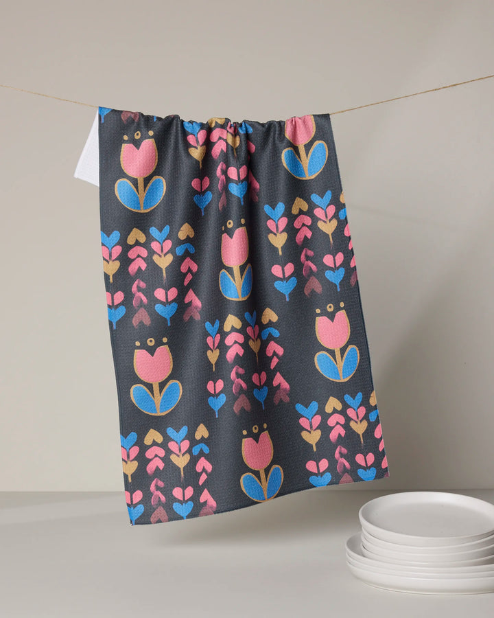 A dark eco-friendly Geometry "Tulip Patch" kitchen towel, featuring a vibrant pink, blue, and gold tulip pattern, hangs on a string above a neat stack of white plates set on a light surface.