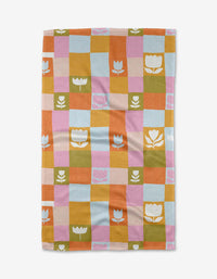 The Tulip Picnic tea towel by Lisa Nelson features a checkered pattern in pastel blue, pink, orange, yellow, and green with white or pink floral and tulip designs. Made from recycled materials, it is shown laid flat on a white background.