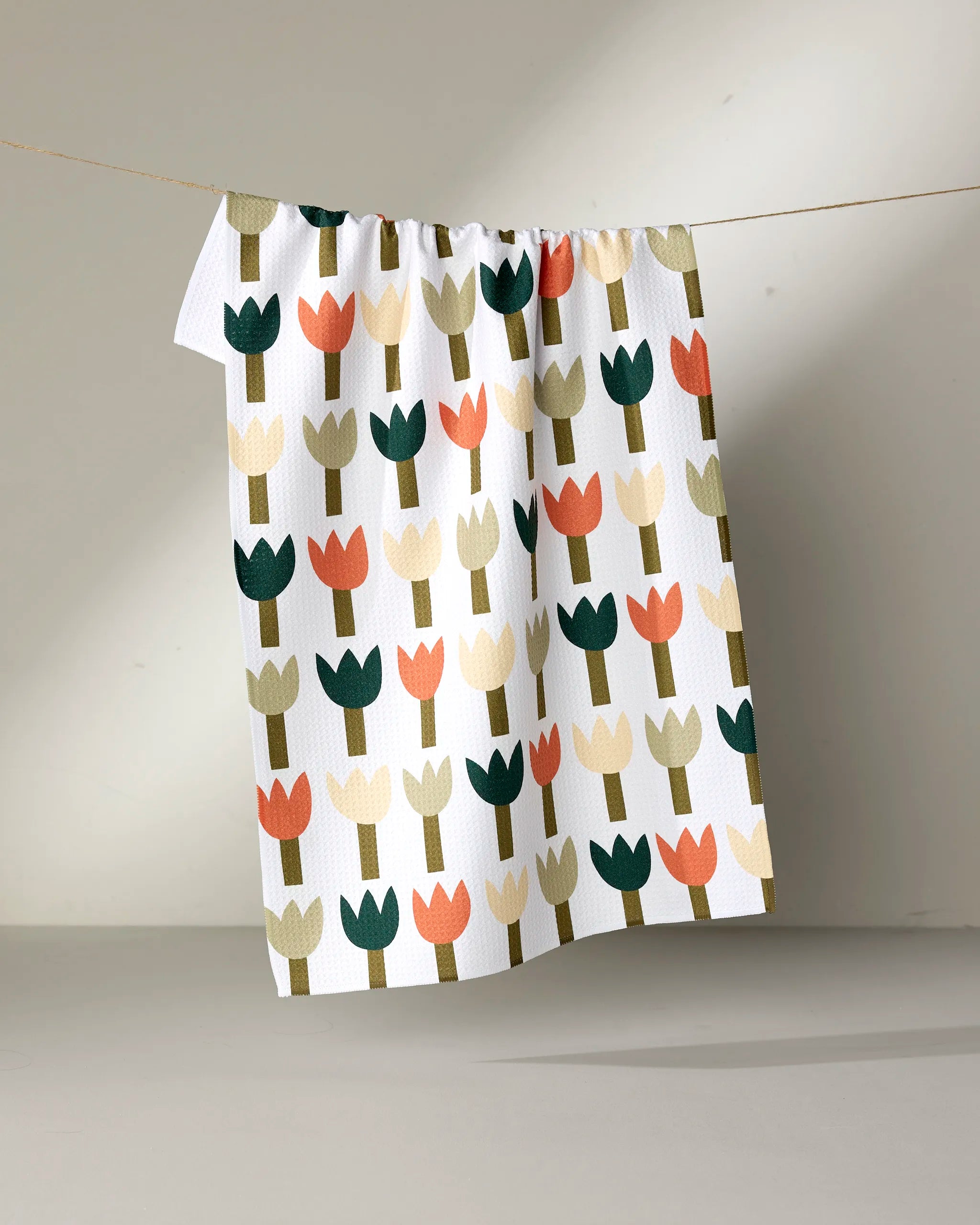 Rachel Parker’s Tulip Row tea towel, made from recycled materials and decorated with stylized tulips in green, peach, beige, and dark blue, hangs against a neutral background.