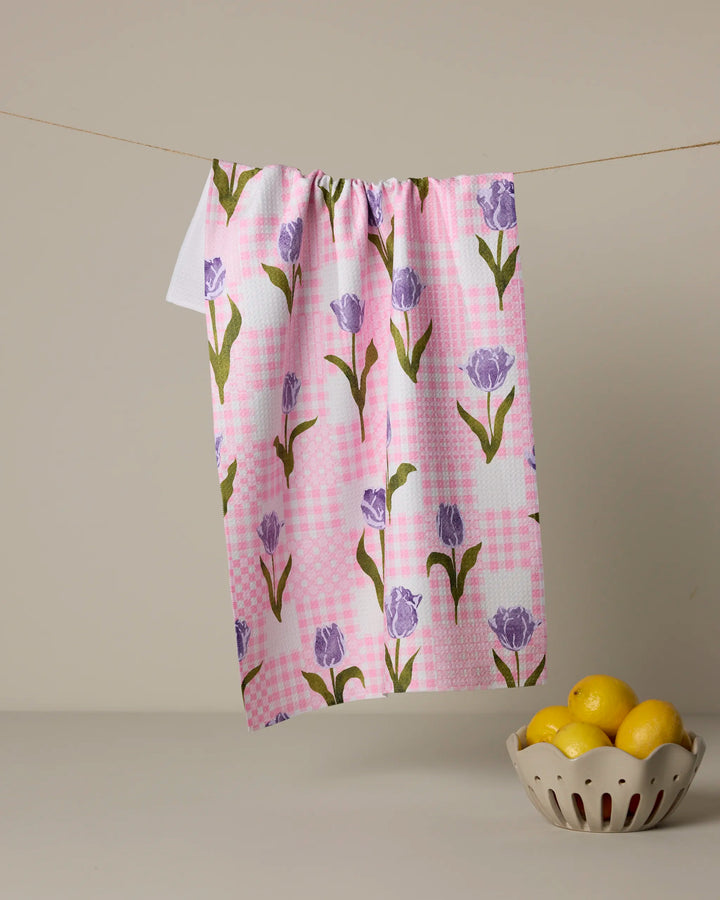 The Geometry Tulip Town tea towel, made from recycled materials and featuring pink, white, and purple tulip designs, hangs on a string above a white bowl of yellow lemons against a beige background.