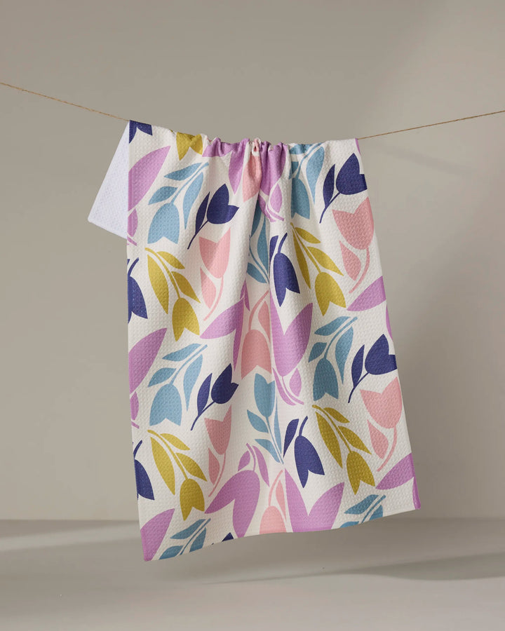 The Lizzie Clark Tulip Tumble is a white, absorbent kitchen towel featuring a colorful leaf pattern in purple, pink, yellow, and blue. It’s pictured hanging on a clothesline against a plain, light background.