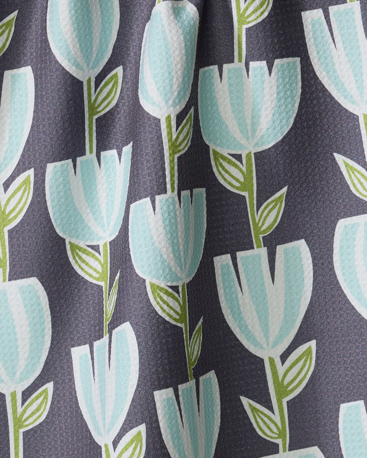 The Lizzie Clark "Tulips for Days" kitchen towel features stylized light blue tulips with green stems and leaves on a dark gray background, crafted from slightly textured fabric with gentle folds.