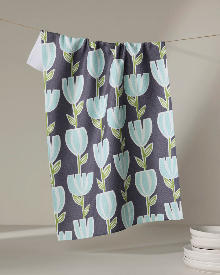 The "Tulips for Days" tea towel by Lizzie Clark, crafted from recycled materials and adorned with blue tulips and green stems on a dark background, hangs on a clothesline beside a stack of white plates.
