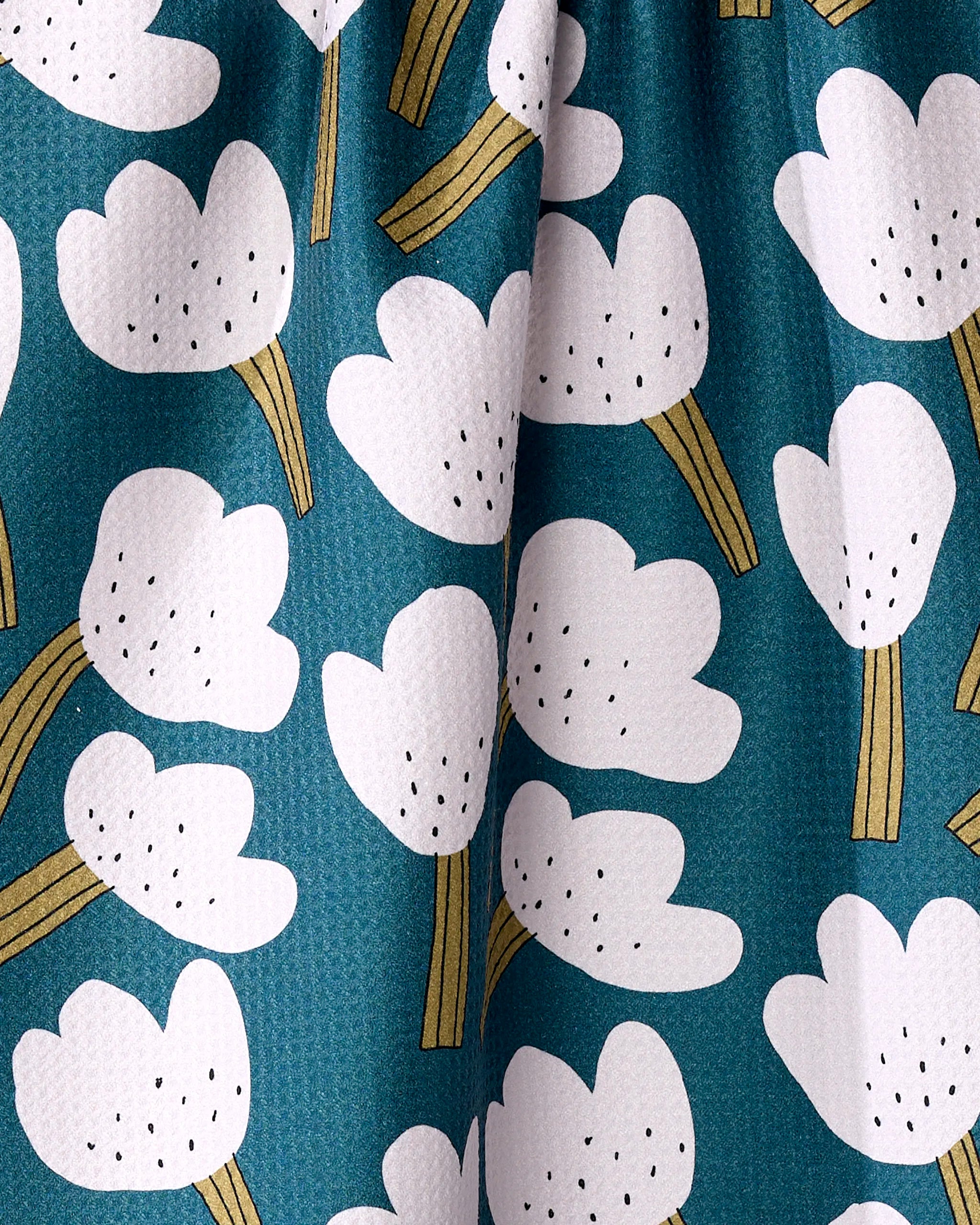 A close-up of the Tulips kitchen towel by Jennifer Bouron, featuring eco-friendly fabric with a repeating pattern of white abstract flowers, yellow stems, and black dots on a teal background. The material appears slightly wrinkled.