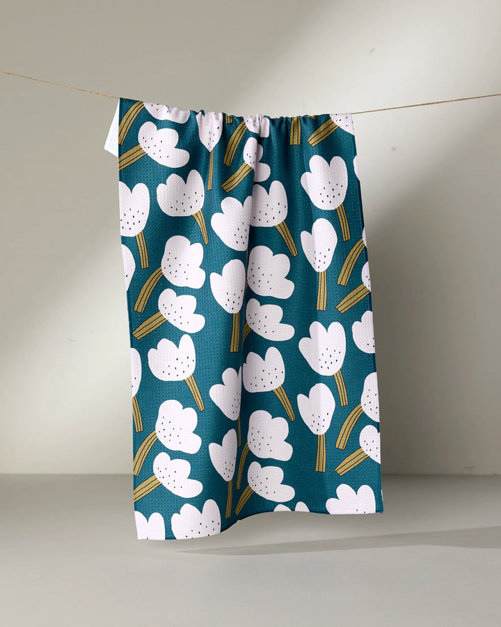 The Tulips tea towel by Jennifer Bouron, in blue with white abstract flowers and yellow stems, hangs on a string against a plain, light background.