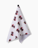The Turkey Trot tea towel by Vaishali Design Studio features a repeating pattern of black and red roosters, hanging from a black loop against a plain white background.