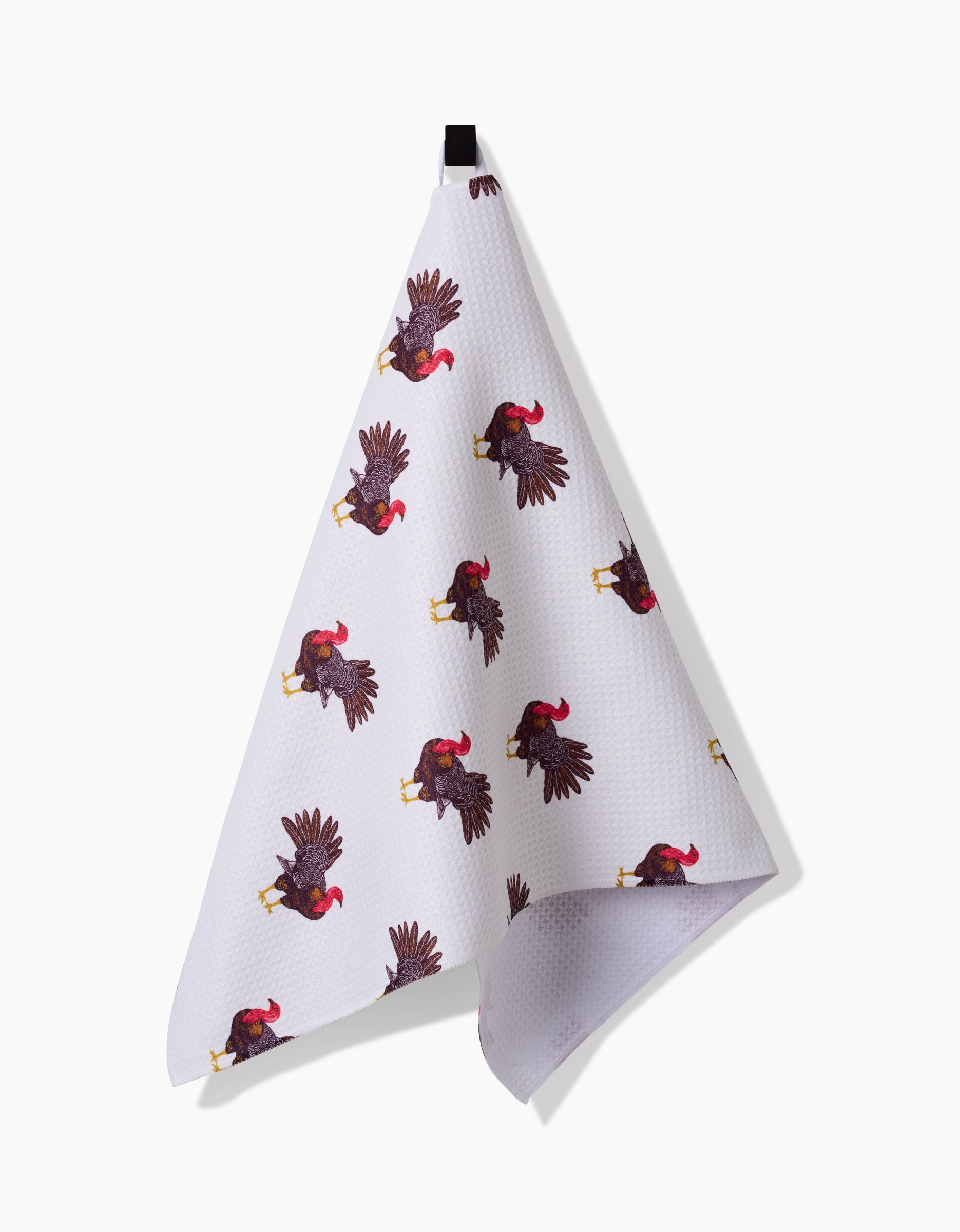 The Turkey Trot tea towel by Vaishali Design Studio features a repeating pattern of black and red roosters, hanging from a black loop against a plain white background.