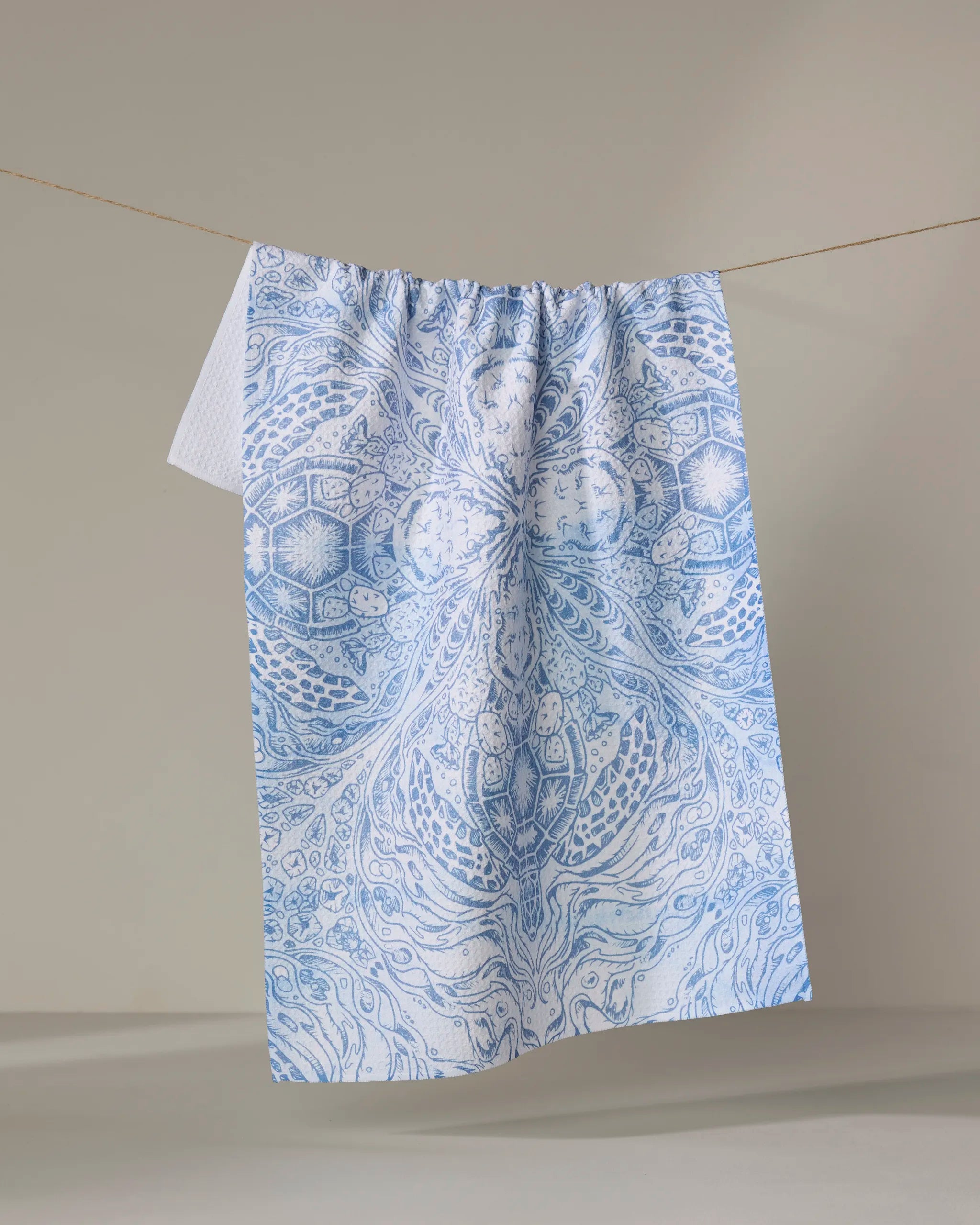 A Skye Walker "Turtle" recycled kitchen towel, featuring blue and white symmetrical patterns, hangs on a clothesline against a neutral background.