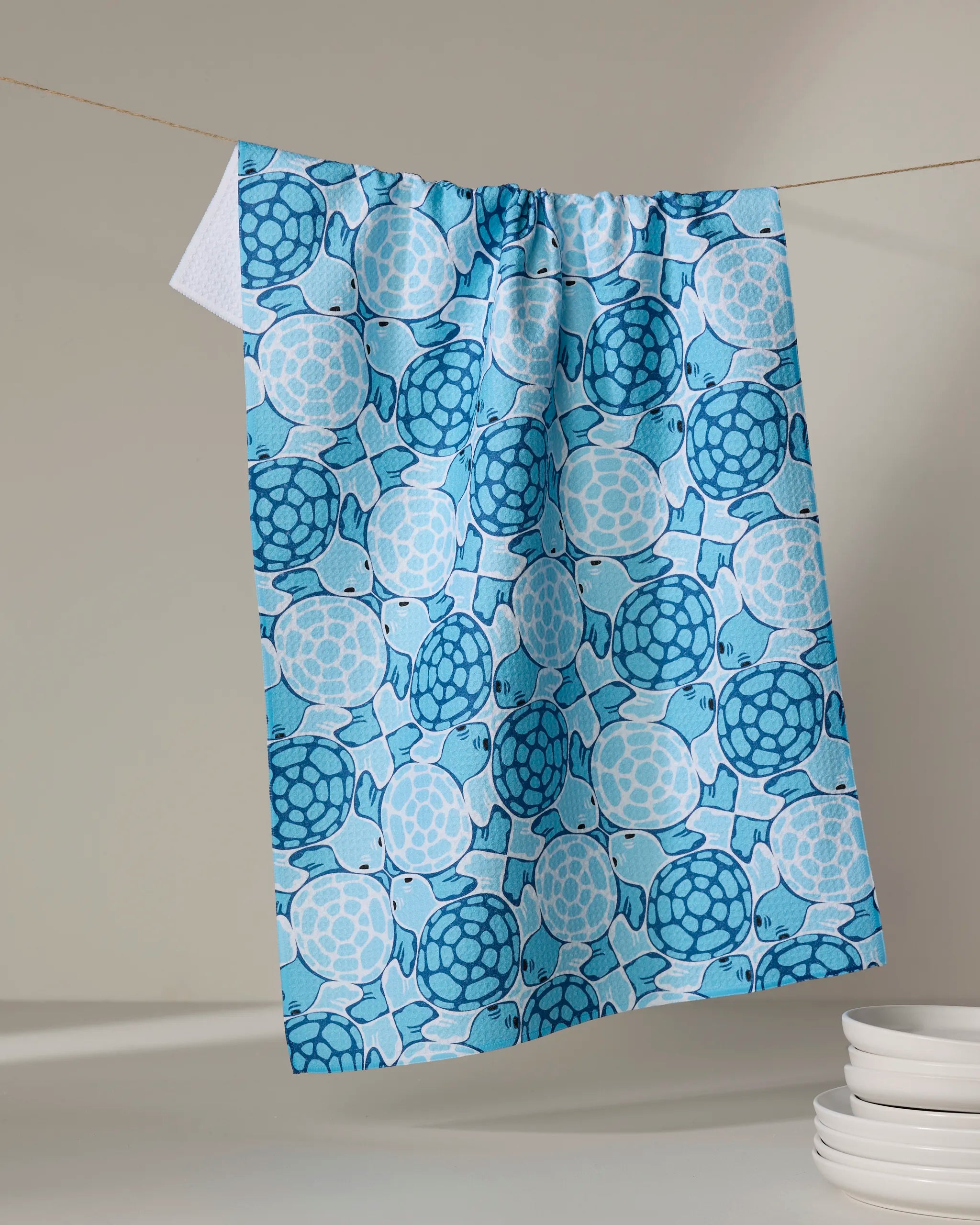 A Marta Harvey "Turtles" blue recycled kitchen towel featuring cartoon sea turtles in various blue shades hangs on a clothesline above stacked white plates.