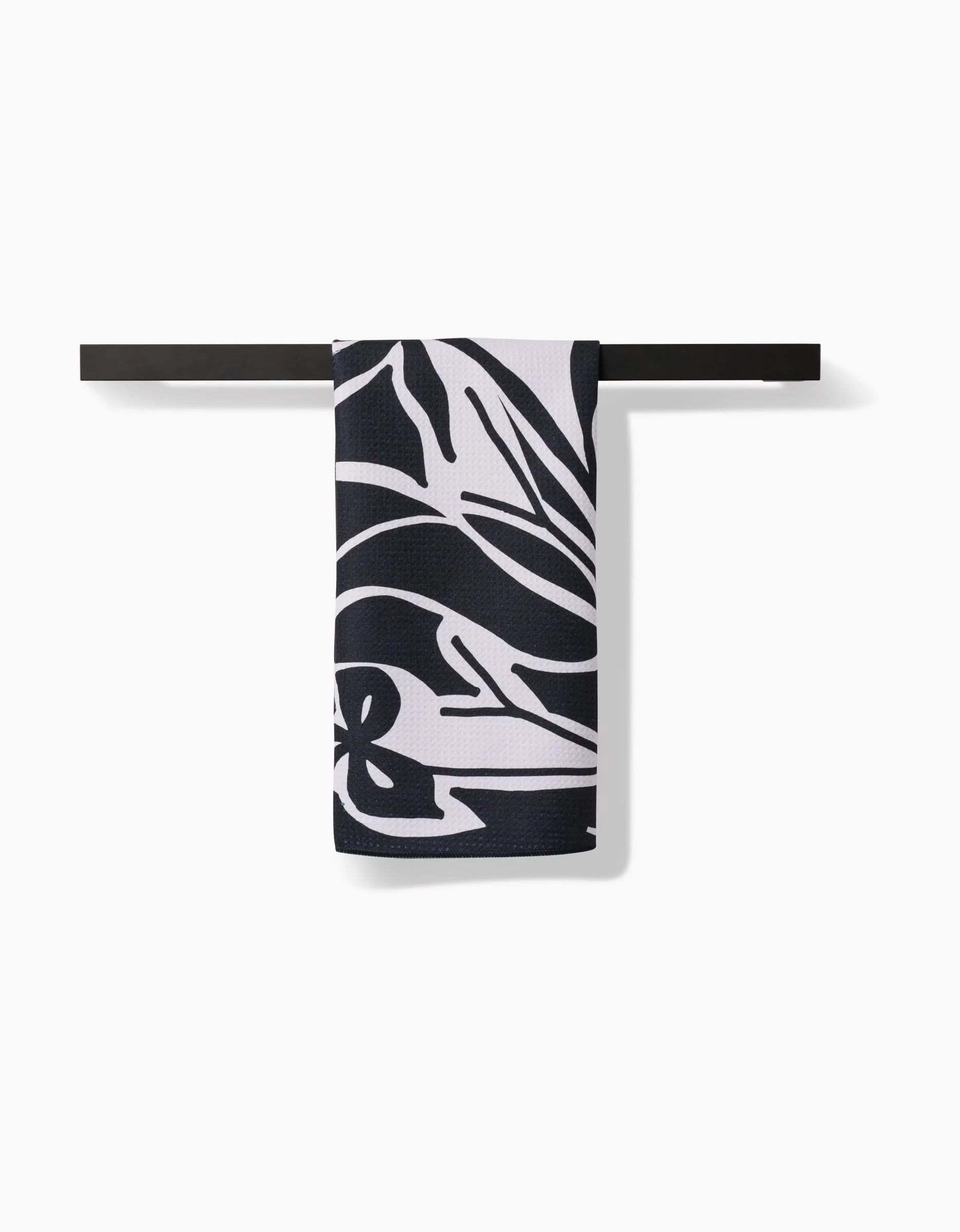 The Ash Cascade Twilight Plant kitchen towel, featuring a bold black and white abstract floral pattern, hangs neatly folded over a sleek black bar against a white background.