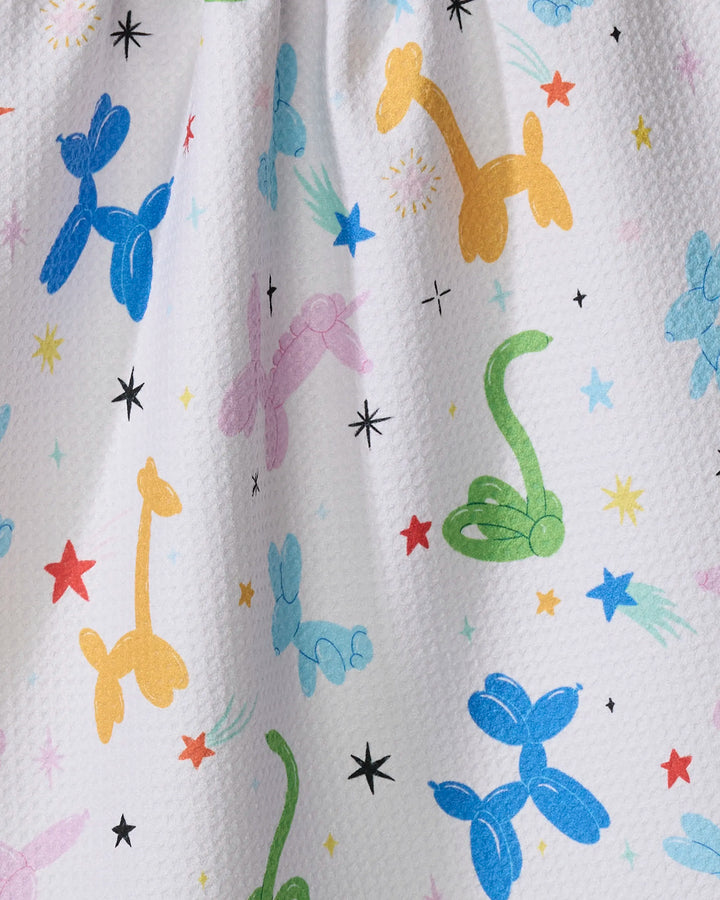 The Twisties tea towel by Vaishali Design Studio features a white textured design with colorful balloon animals—giraffes, dogs, and snakes—in blue, yellow, green, and pink, scattered with multicolored stars and starbursts.