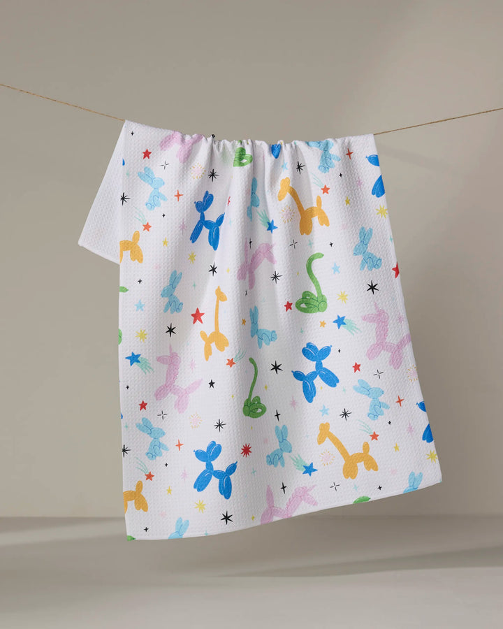 The Twisties tea towel by Vaishali Design Studio dries quickly and features colorful balloon dogs, giraffes, and dinosaurs with small black stars, shown hanging on a clothesline against a neutral background.