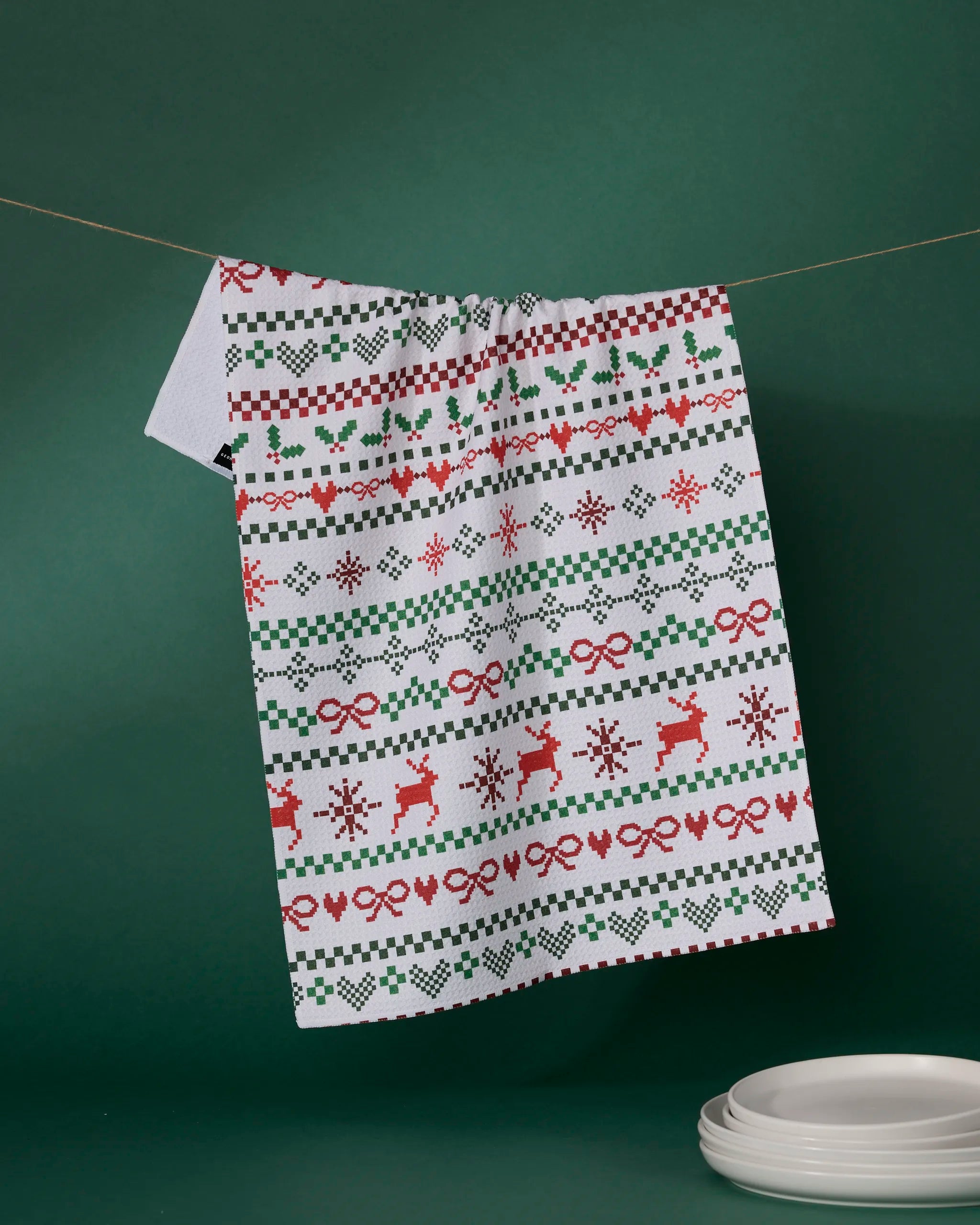 The "Ugly Sweater Day" kitchen towel by Geometry, made from recycled materials and adorned with red and green holiday motifs, hangs in front of a green background, with a stack of white plates nearby.