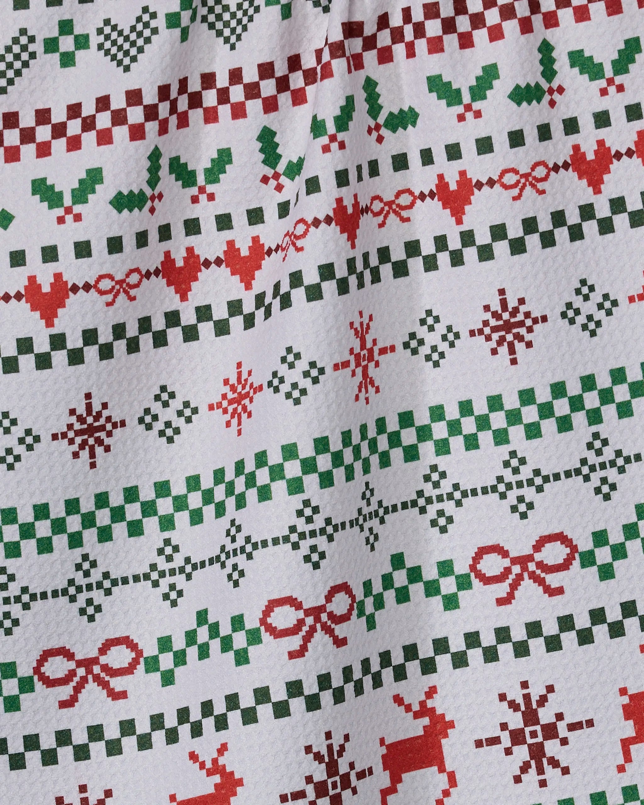 A close-up of the Geometry "Ugly Sweater Day" kitchen towel, showcasing a festive pattern with red and green shapes, holly leaves, stars, bows, and reindeer in a classic holiday sweater style.