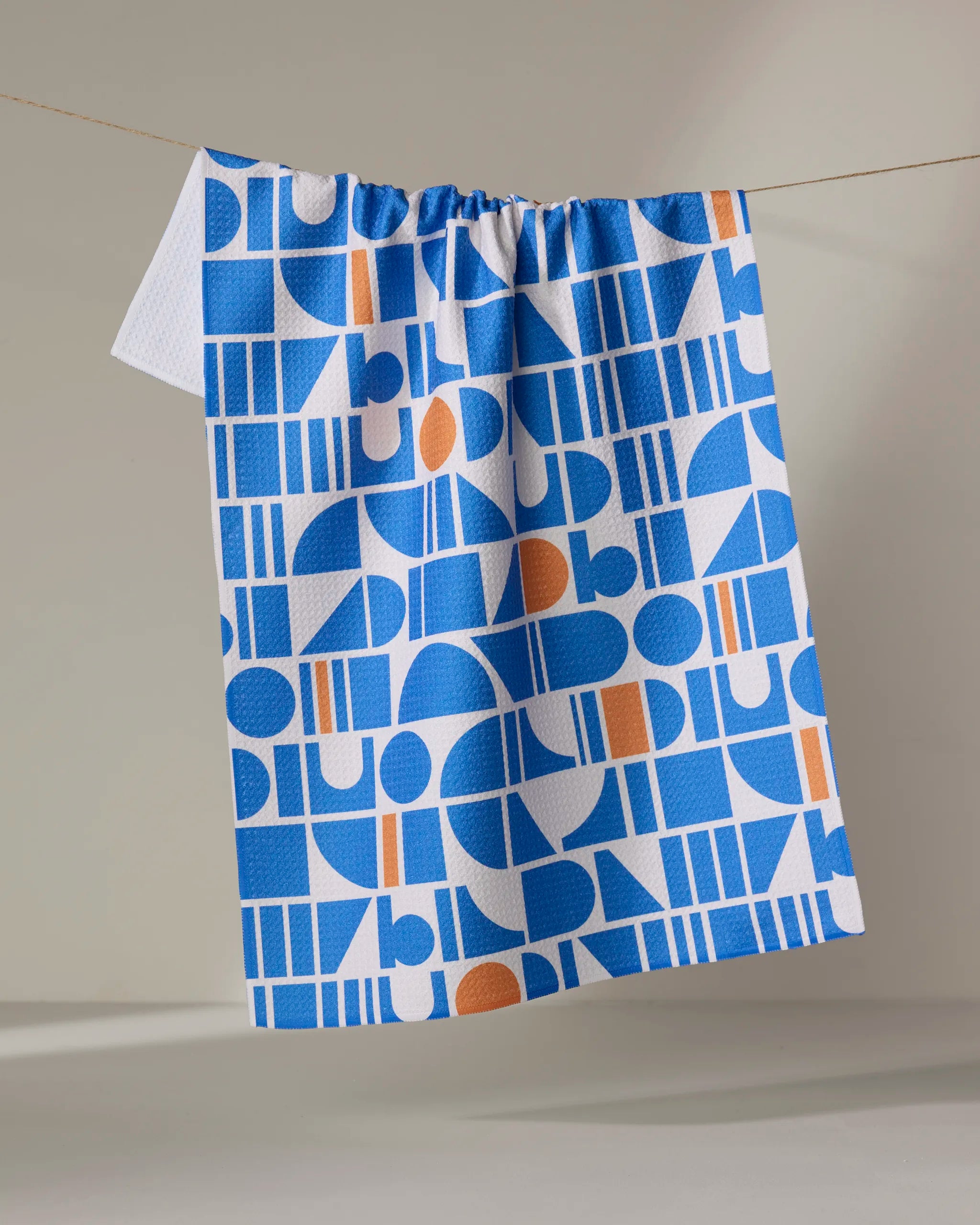 The Happy Hemera Ultra Marine Harmony tea towel, featuring blue and white geometric shapes with orange accents, hangs on a line against a neutral background.