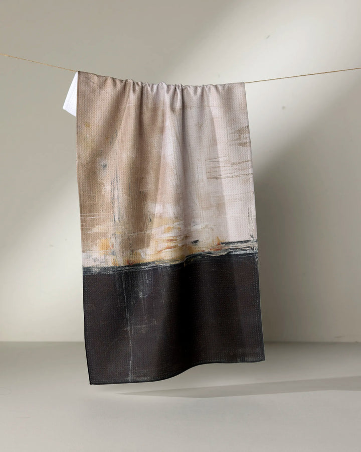 The "Uncharted Territory" tea towel by Jennifer Bouron, featuring an abstract beige, cream, and black design, hangs on a string against a plain light background.