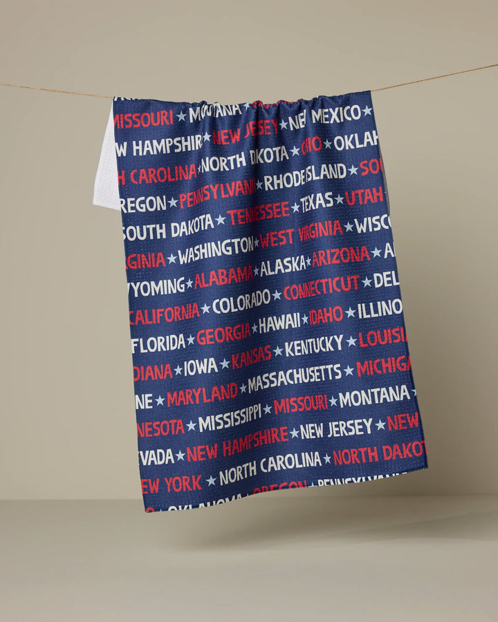 The Geometry "United" kitchen tea towel hangs on a line, featuring the names of all U.S. states in red, white, and blue capital letters on a dark blue background.