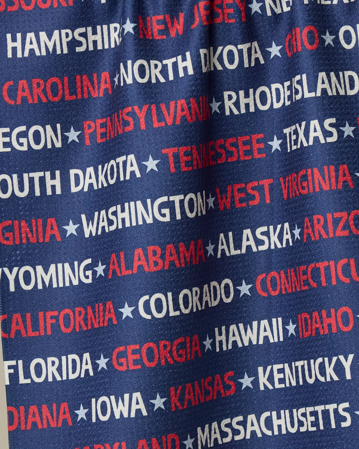 The “United” tea towel by Geometry features U.S. state names in bold white and red capitals on blue, arranged in various directions with stars, and is crafted from recycled materials for eco-friendly appeal.