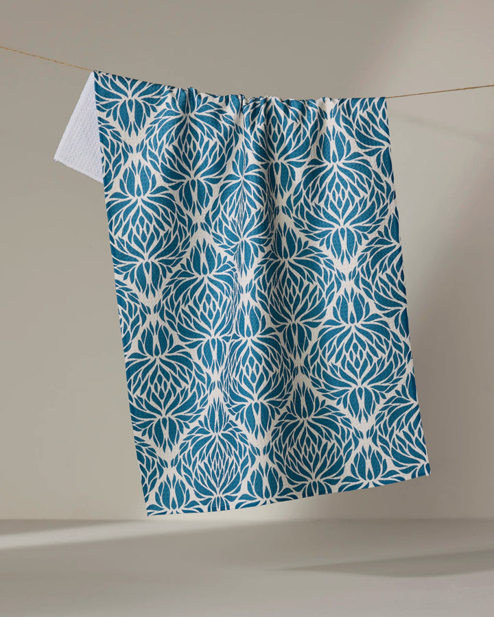 The Untangled tea towel by Rebecca Woolbright features a blue and white lotus-inspired pattern, crafted from recycled materials, and hangs on a clothesline against a plain beige background.